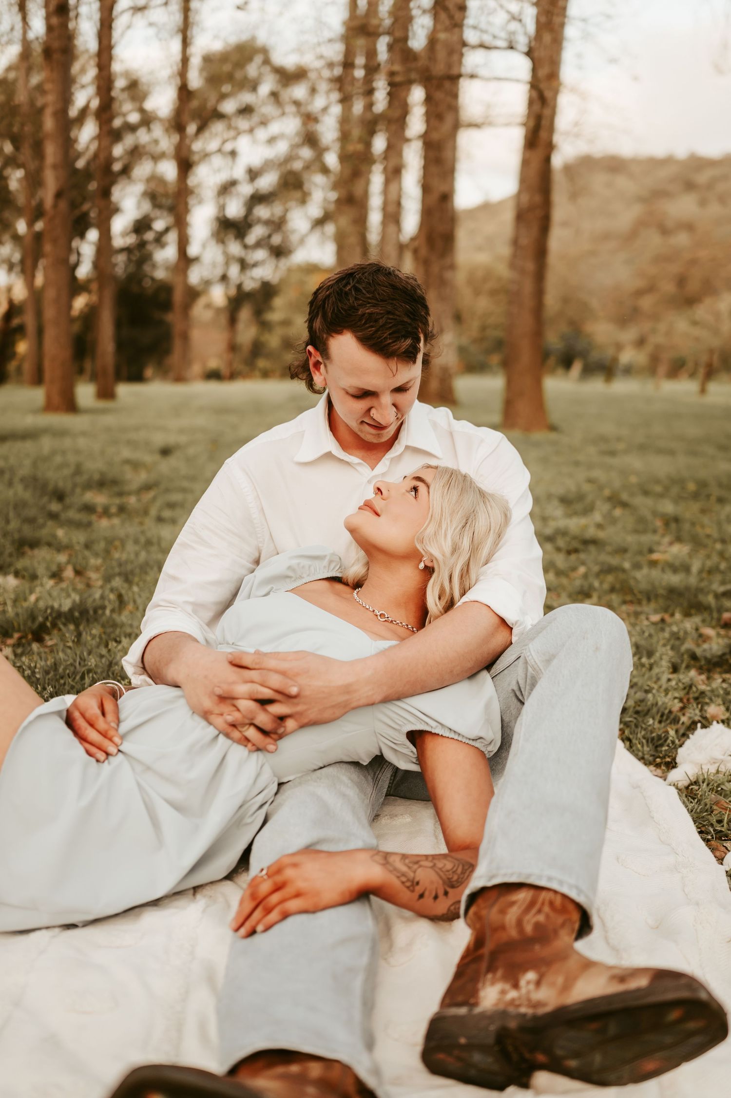 Couple in white clothing shares tender moments while lying in a grassy field among pine trees.