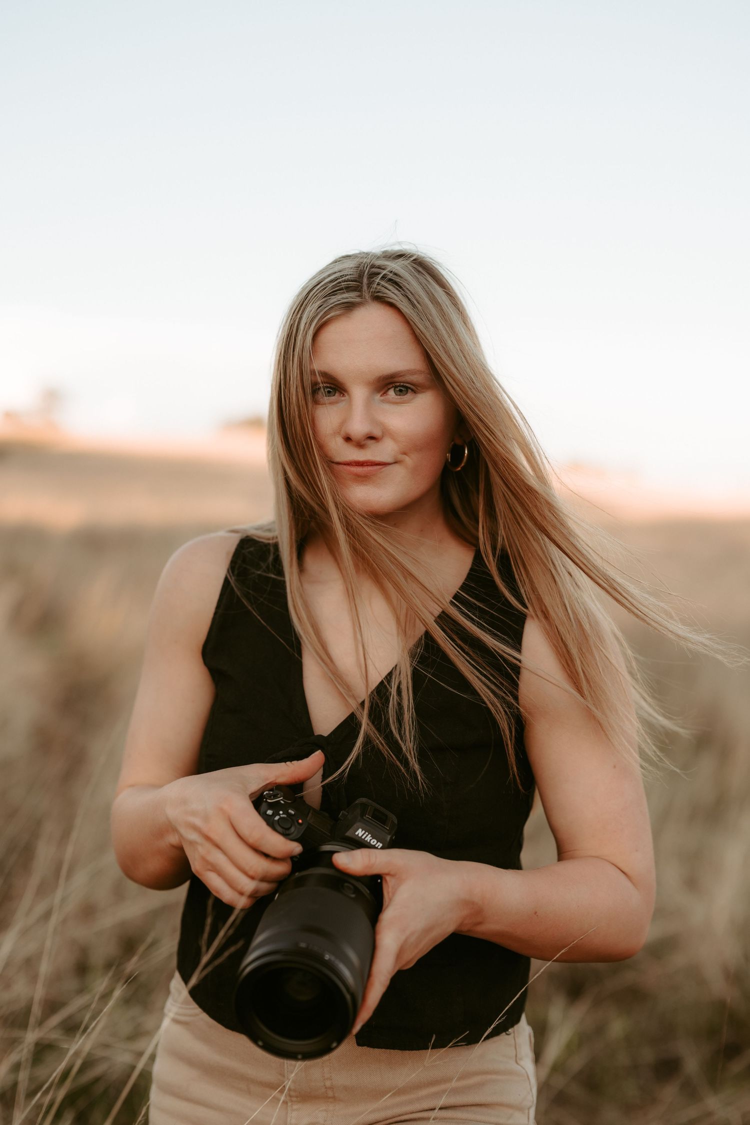 Photographer in black top holding professional camera in golden field at sunset.