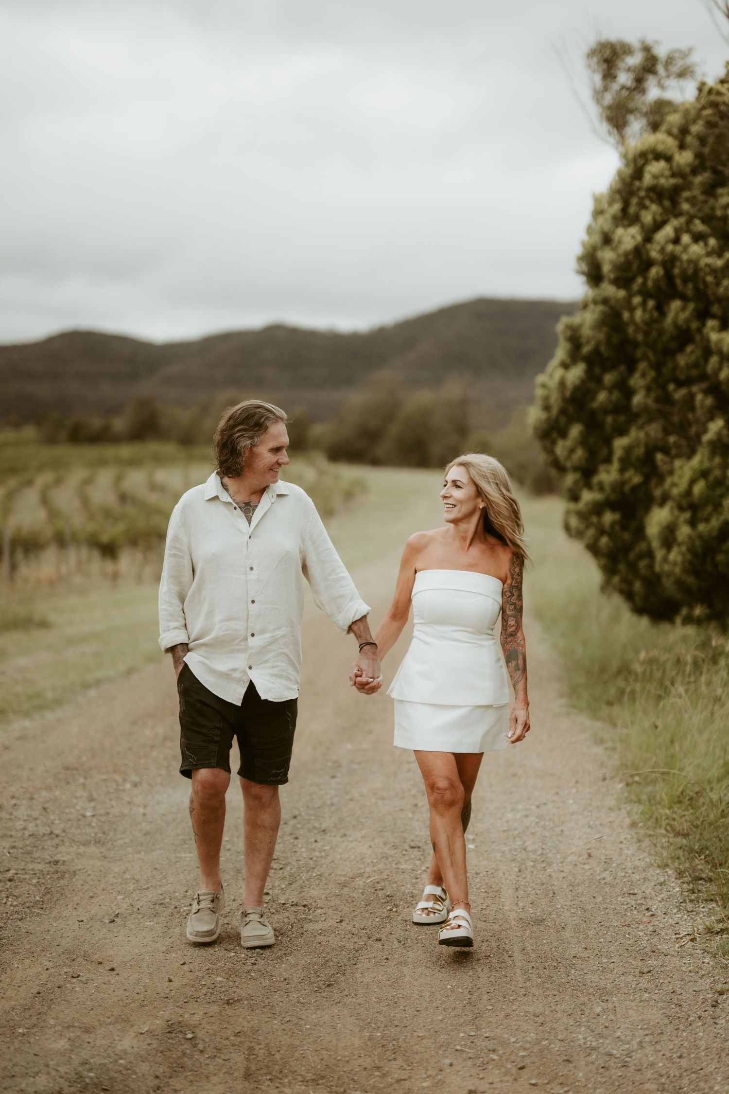 Couple walking hand in hand on a dirt path surrounded by scenic countryside.