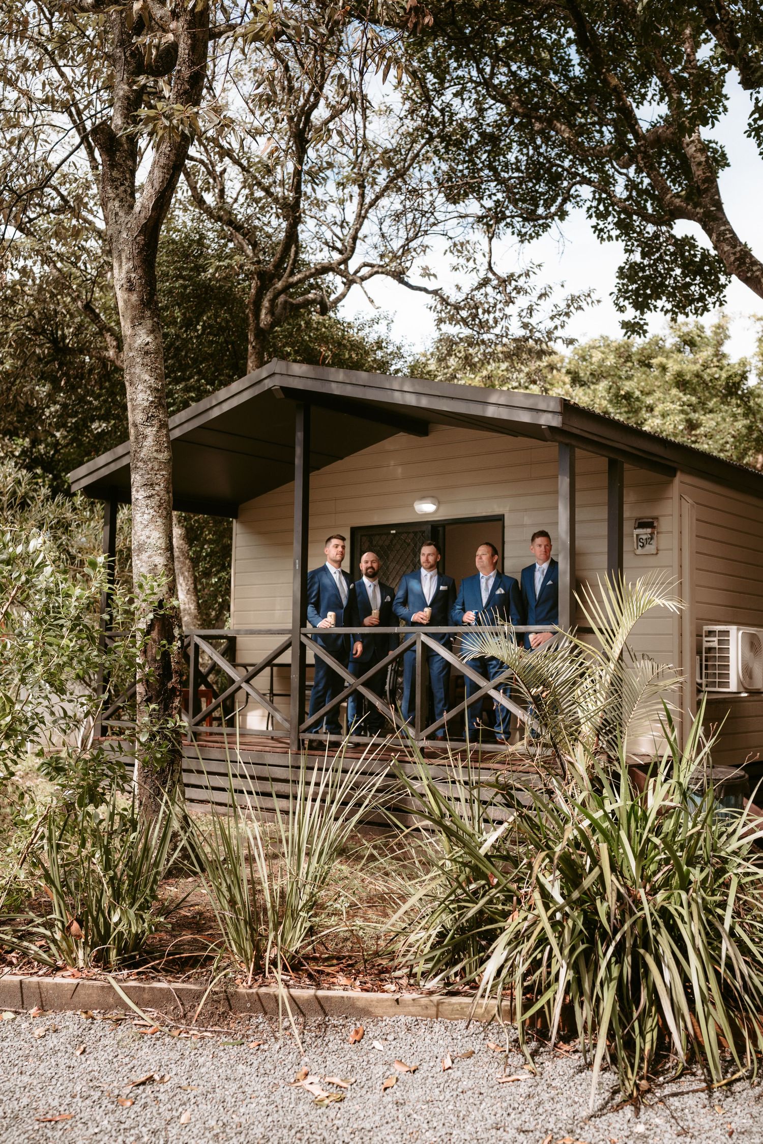 Groomsmen in blue suits pose on the deck of a rustic cabin surrounded by tropical plants.