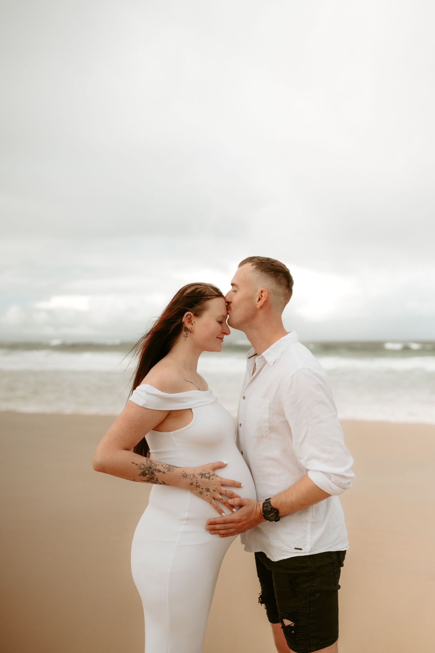 A loving couple embraces on a cloudy beach during their maternity session.
