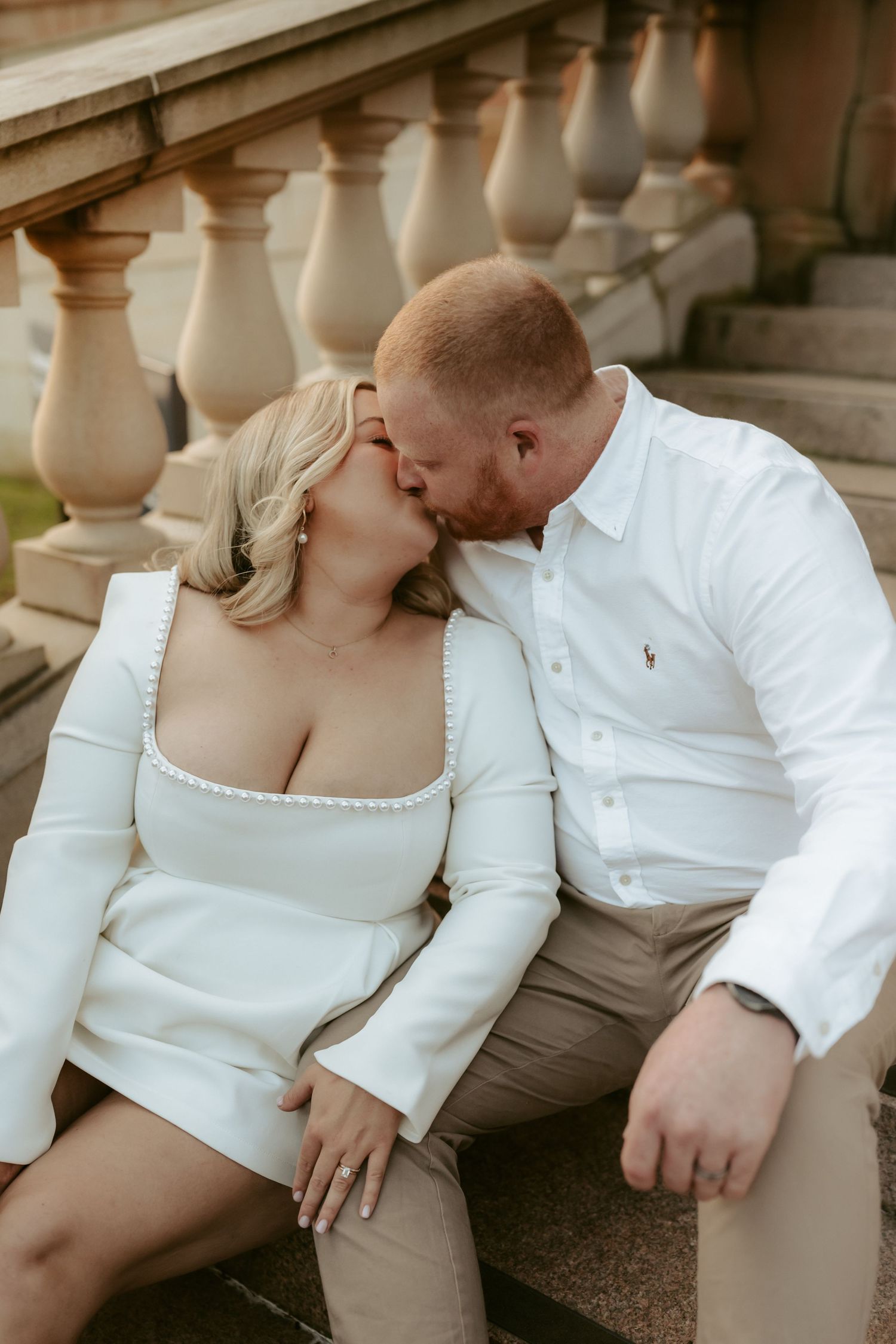 A romantic couple in white and beige outfits share intimate moments on stone steps during a photoshoot.