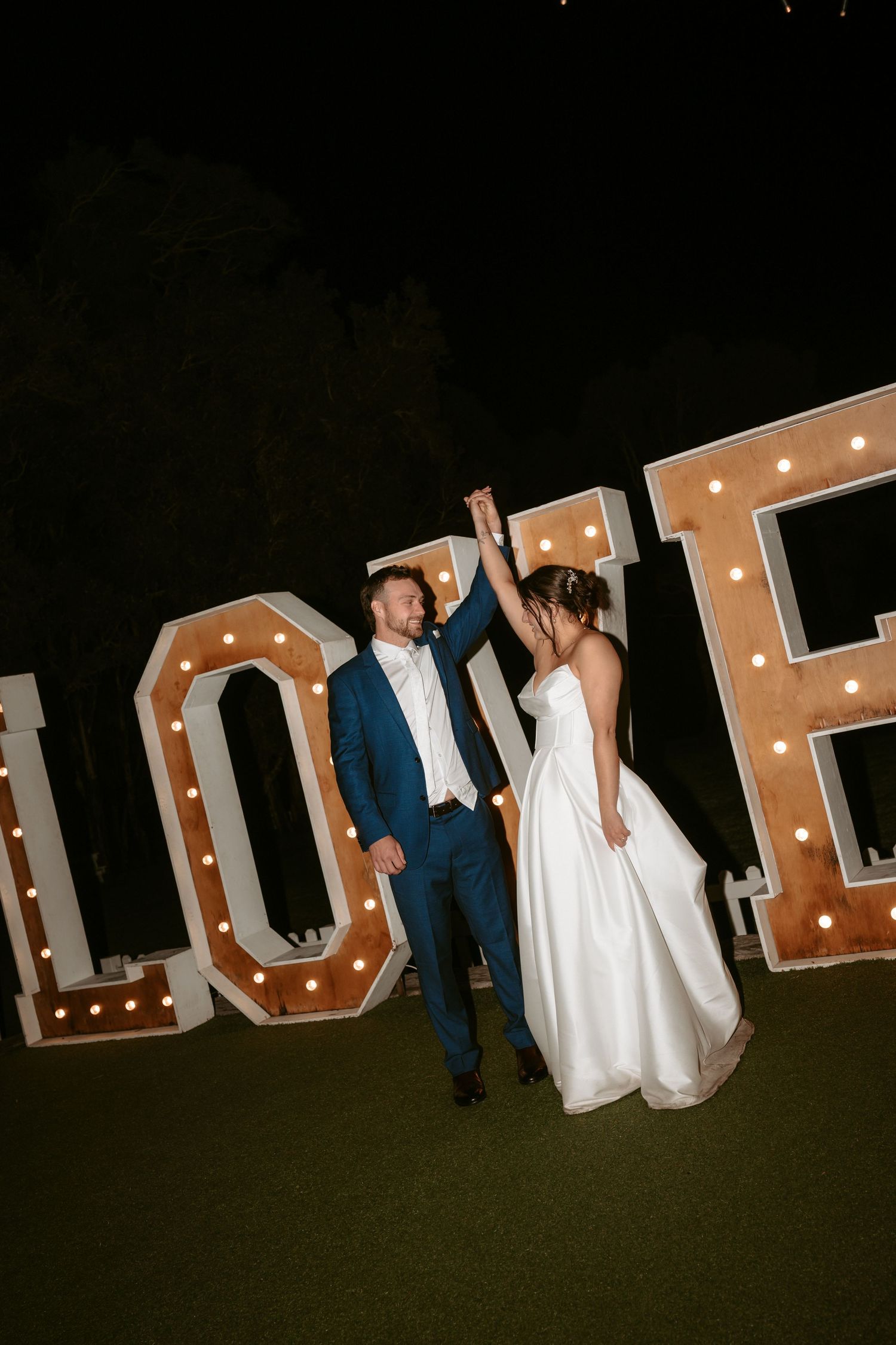 Couple poses in front of illuminated marquee letters at their nighttime wedding celebration.