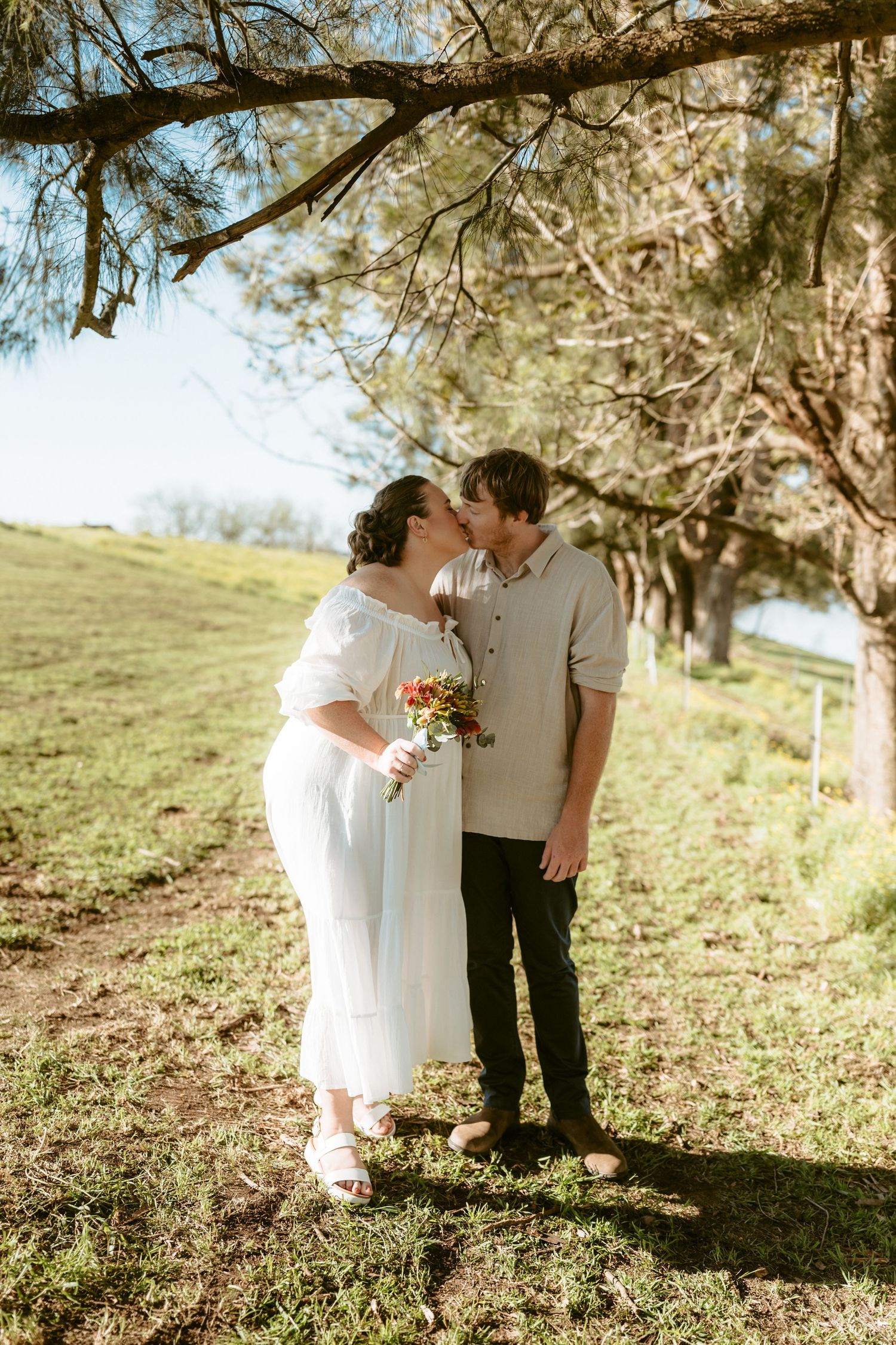 A romantic couple embraces under a tree on a sunny hillside in vintage-style wedding attire.
