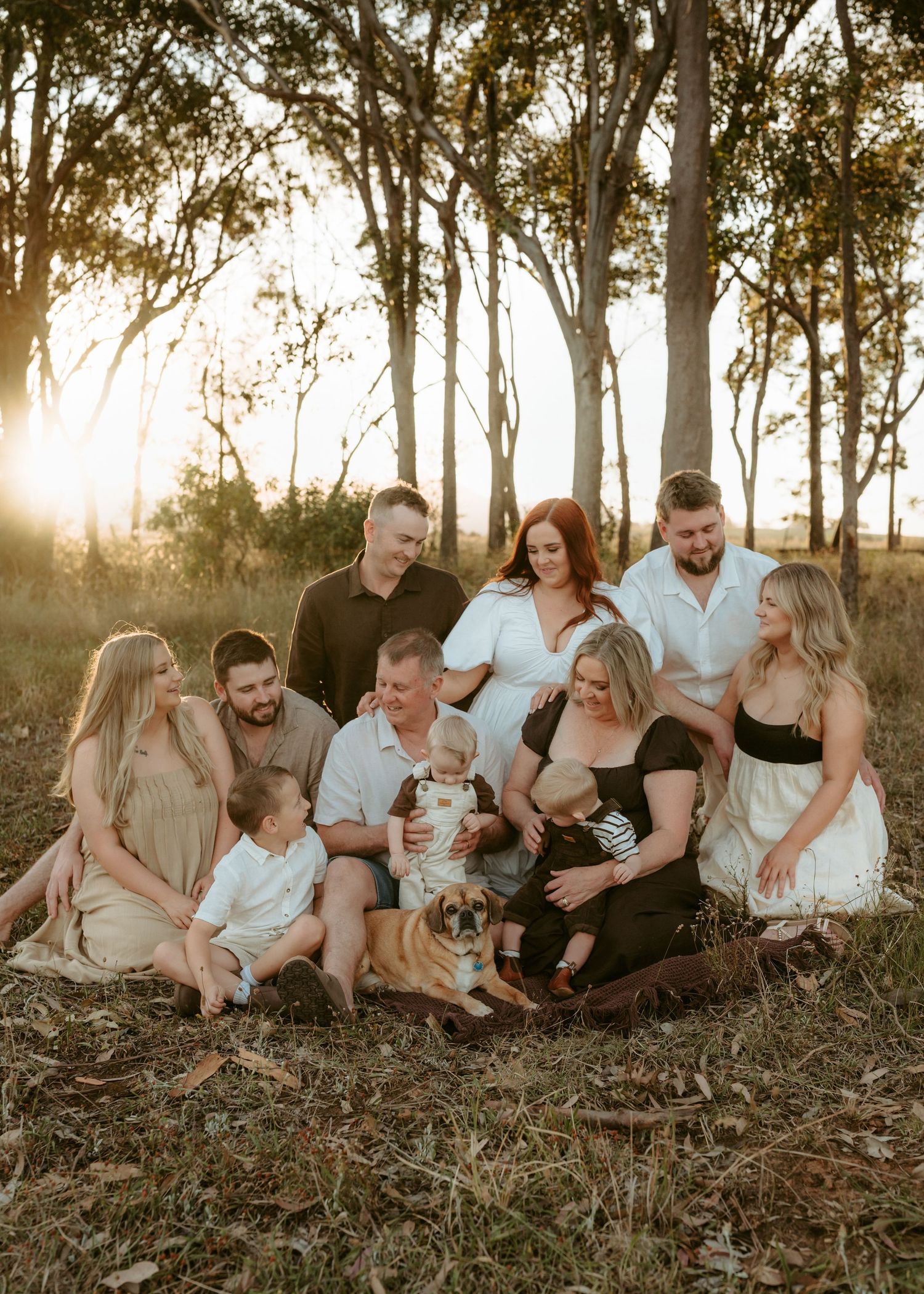 A family sits together on grassy ground during golden hour as sunlight filters through trees in the background.