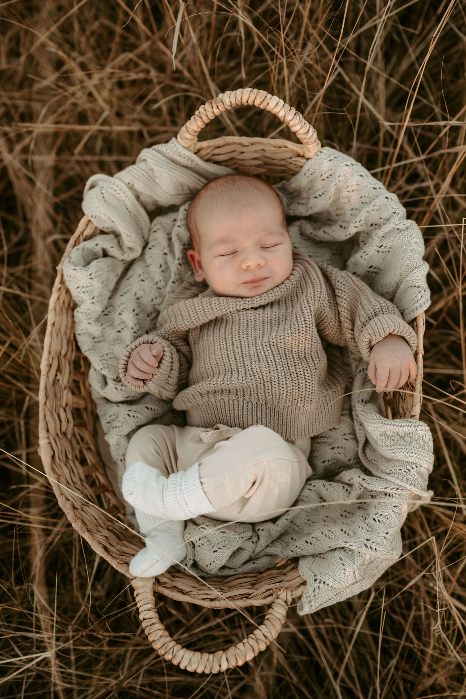 Sleeping newborn in knit sweater nestled in woven basket with soft textiles.