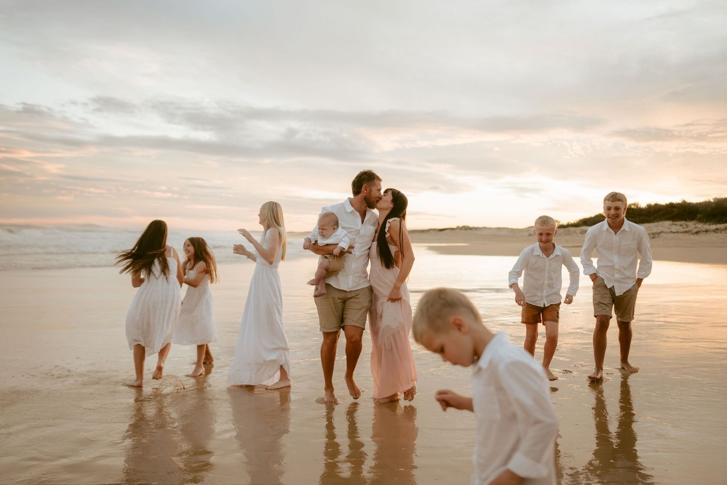 People holding hands and walking along the shoreline at sunset in coordinated white and beige outfits.