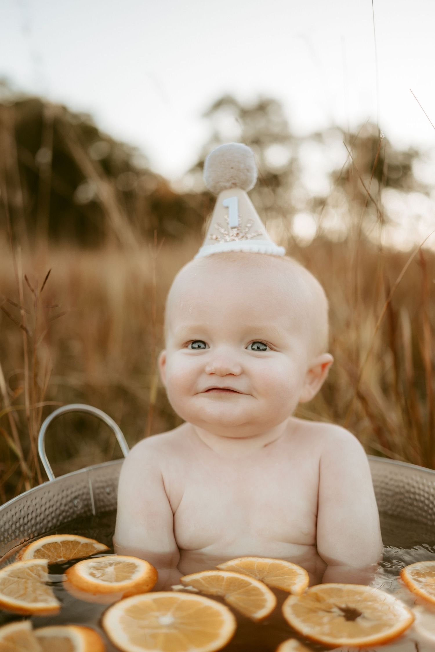 Baby sits in metal basin surrounded by orange slices during outdoor portrait session.