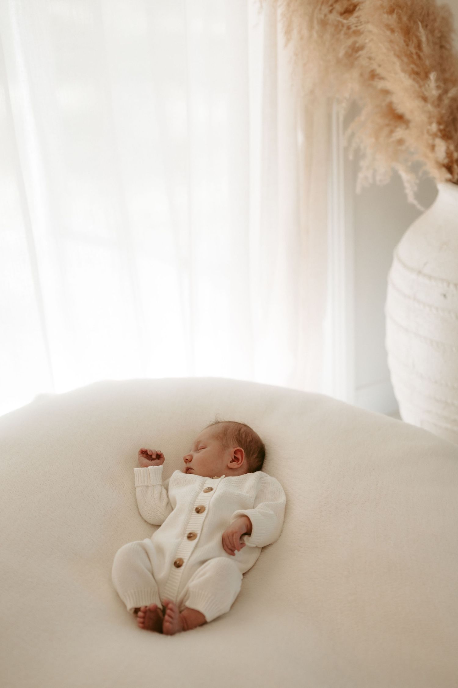 Baby in white outfit lying peacefully on white bedding in natural window light.