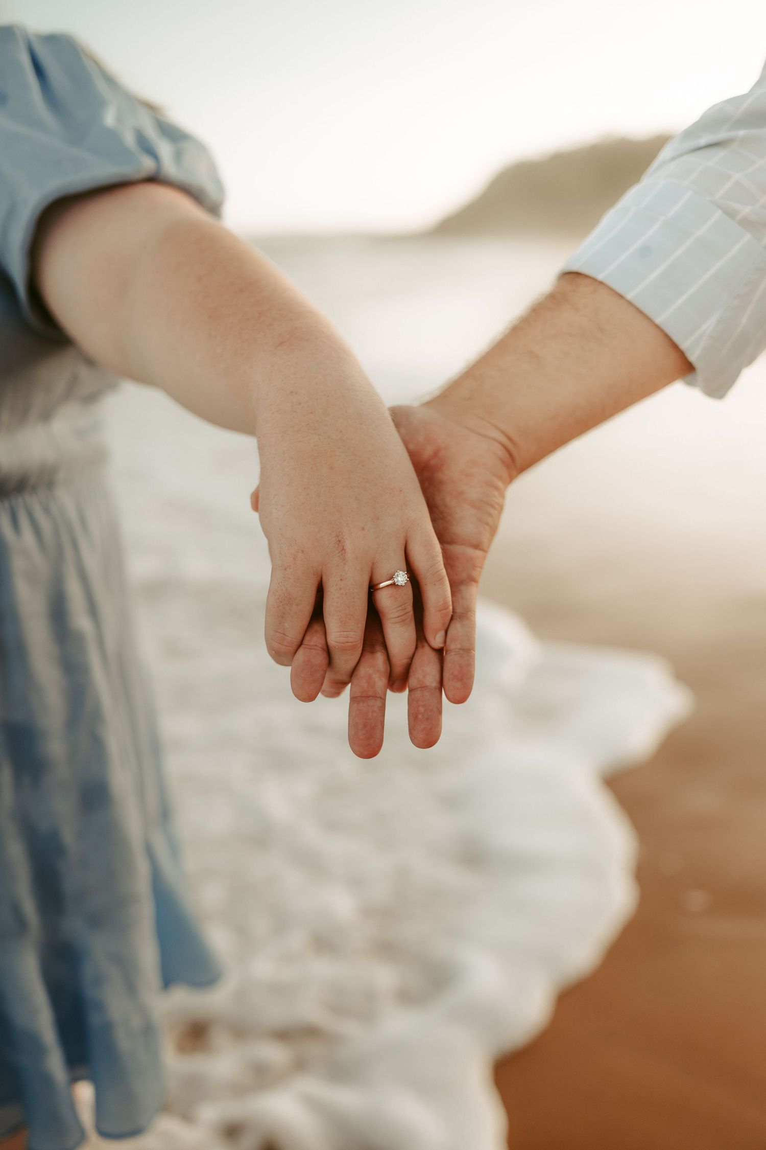 Two hands tenderly clasped together with a wedding ring visible against a soft beach backdrop.