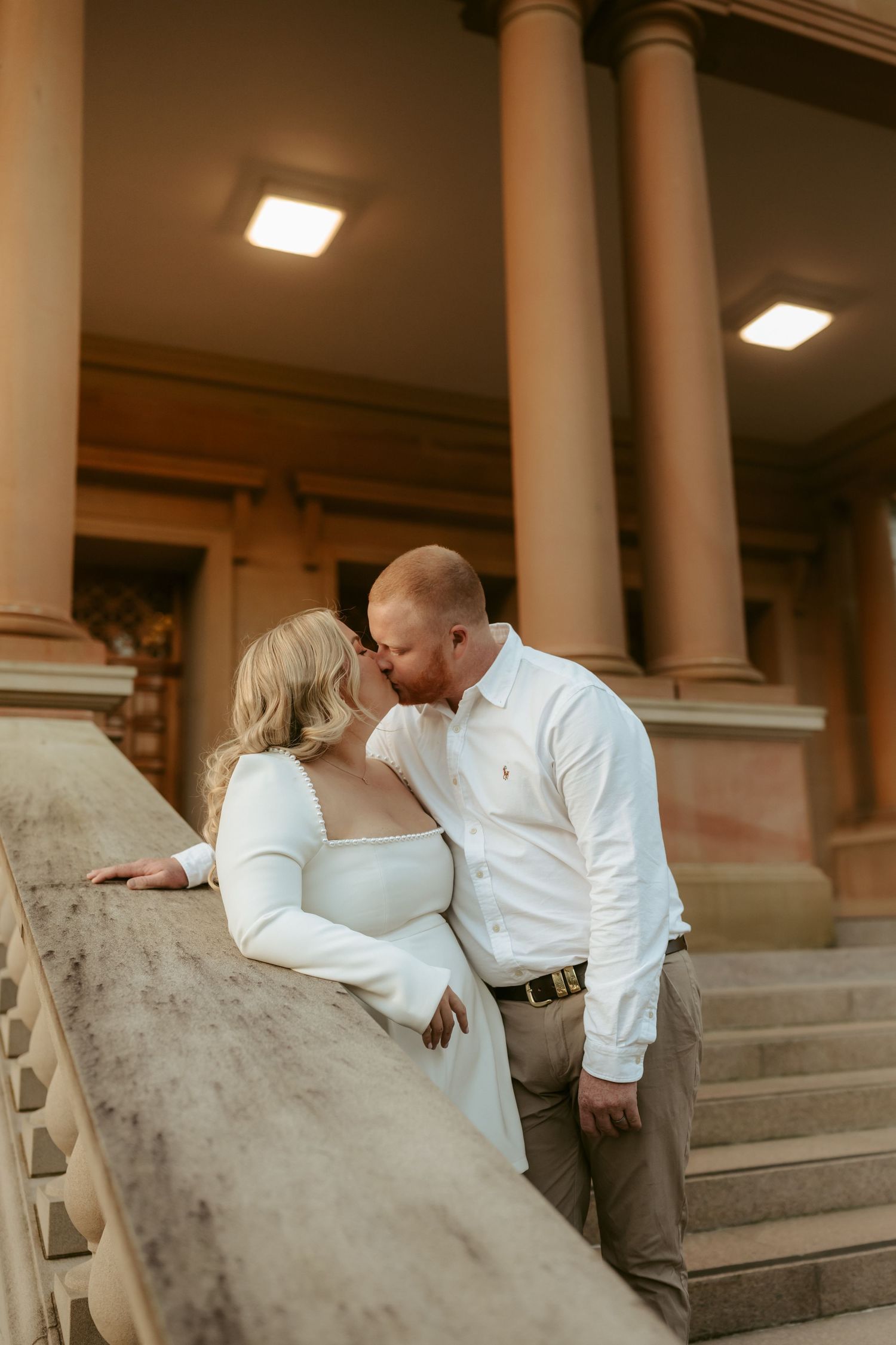 A romantic couple in matching white shirts share an intimate moment on stone steps near elegant columns.