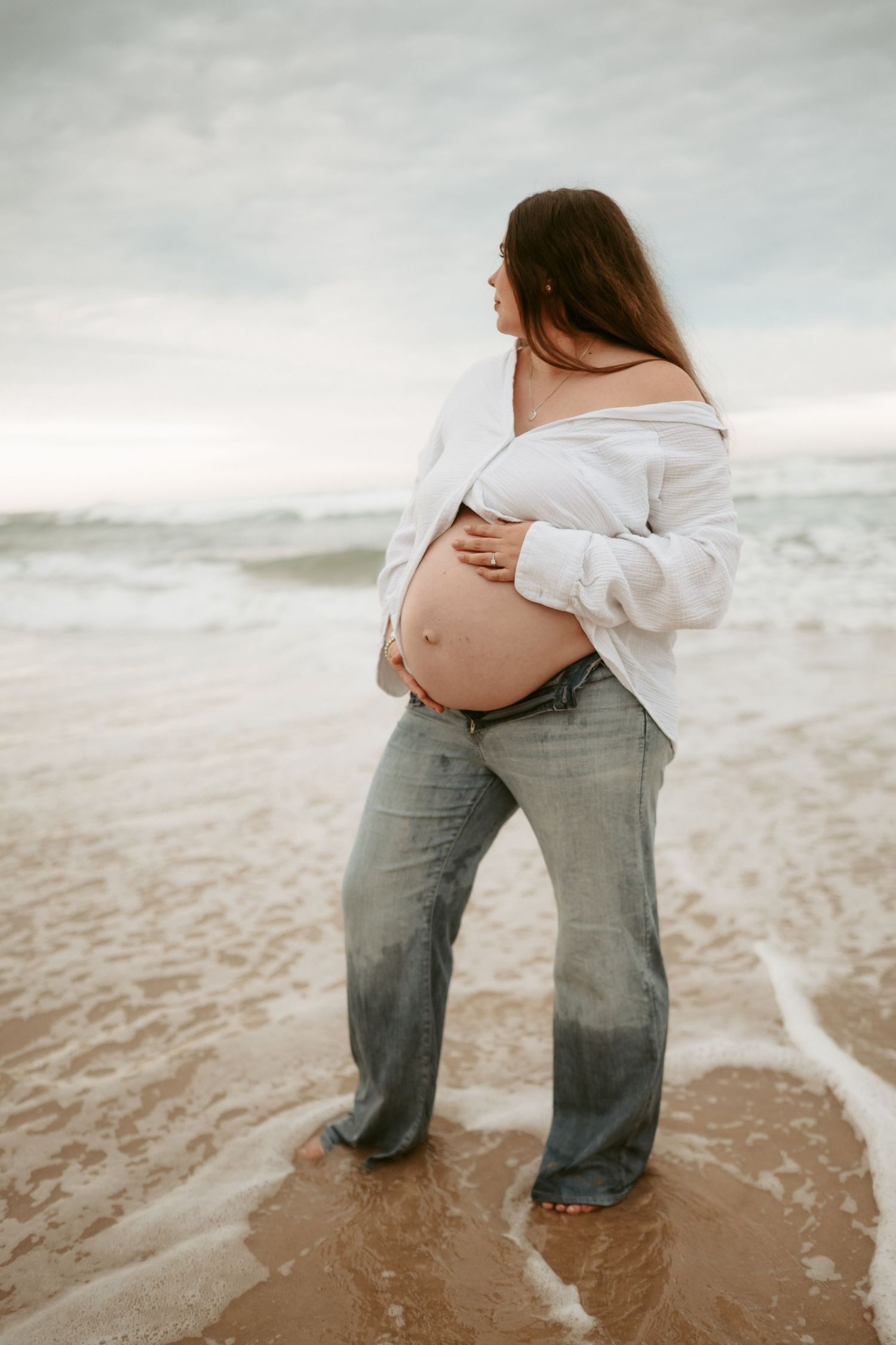 A pregnant woman in a white un-buttoned shirt stands at the ocean's edge during a maternity photoshoot.