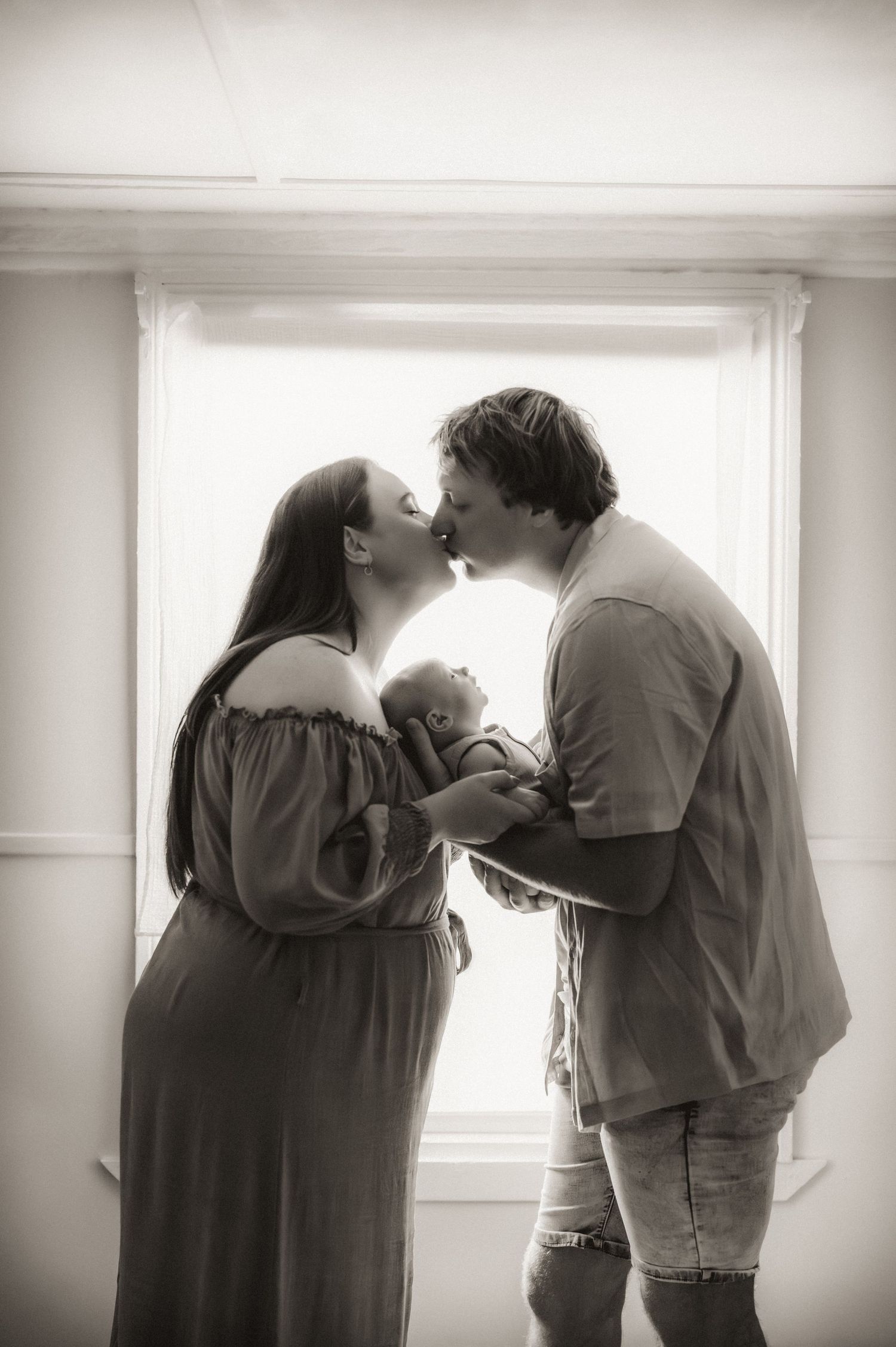 An intimate black and white portrait of parents sharing a kiss while holding their newborn.