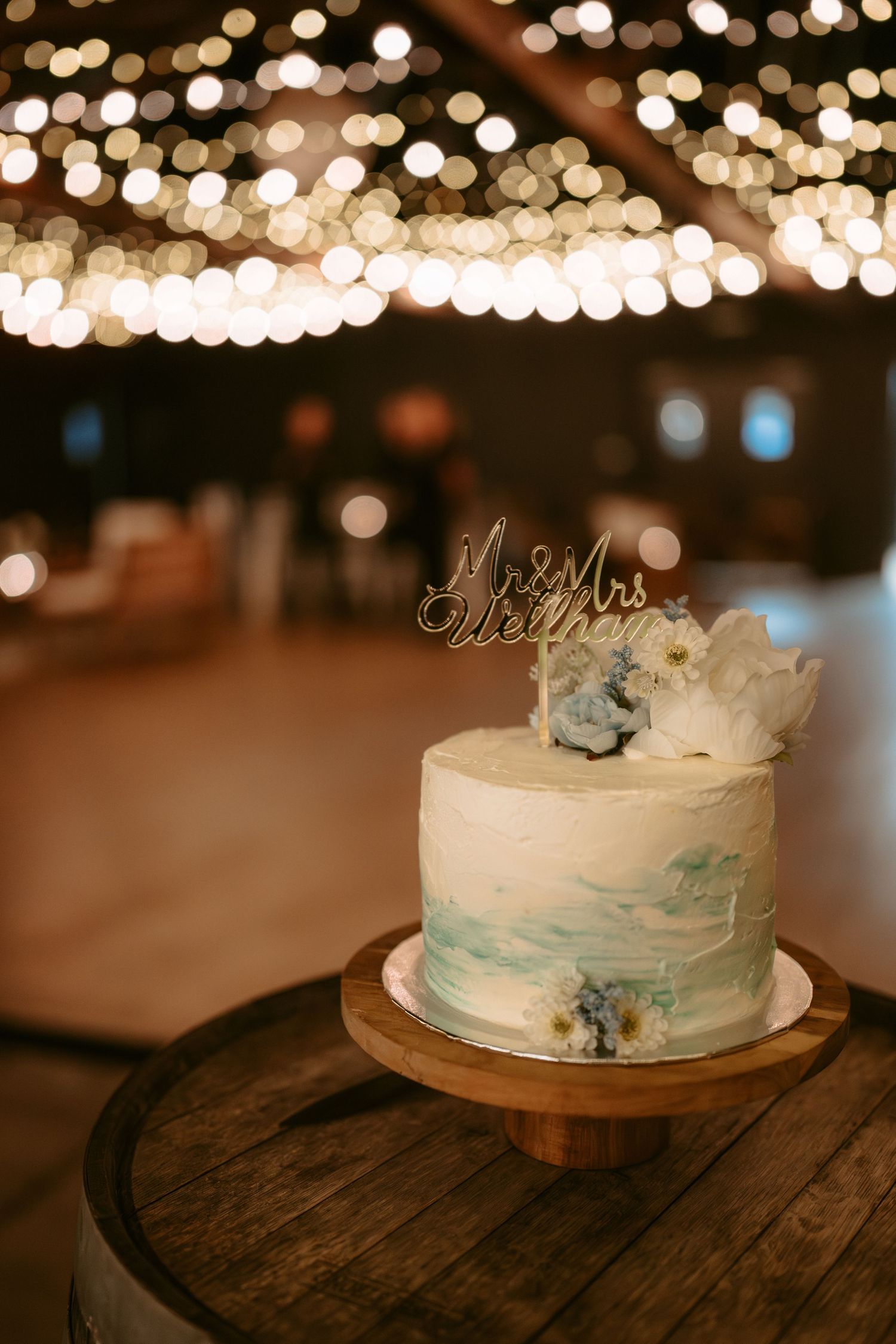 A white wedding cake with blue marbled details sits on a wooden surface under twinkling lights.