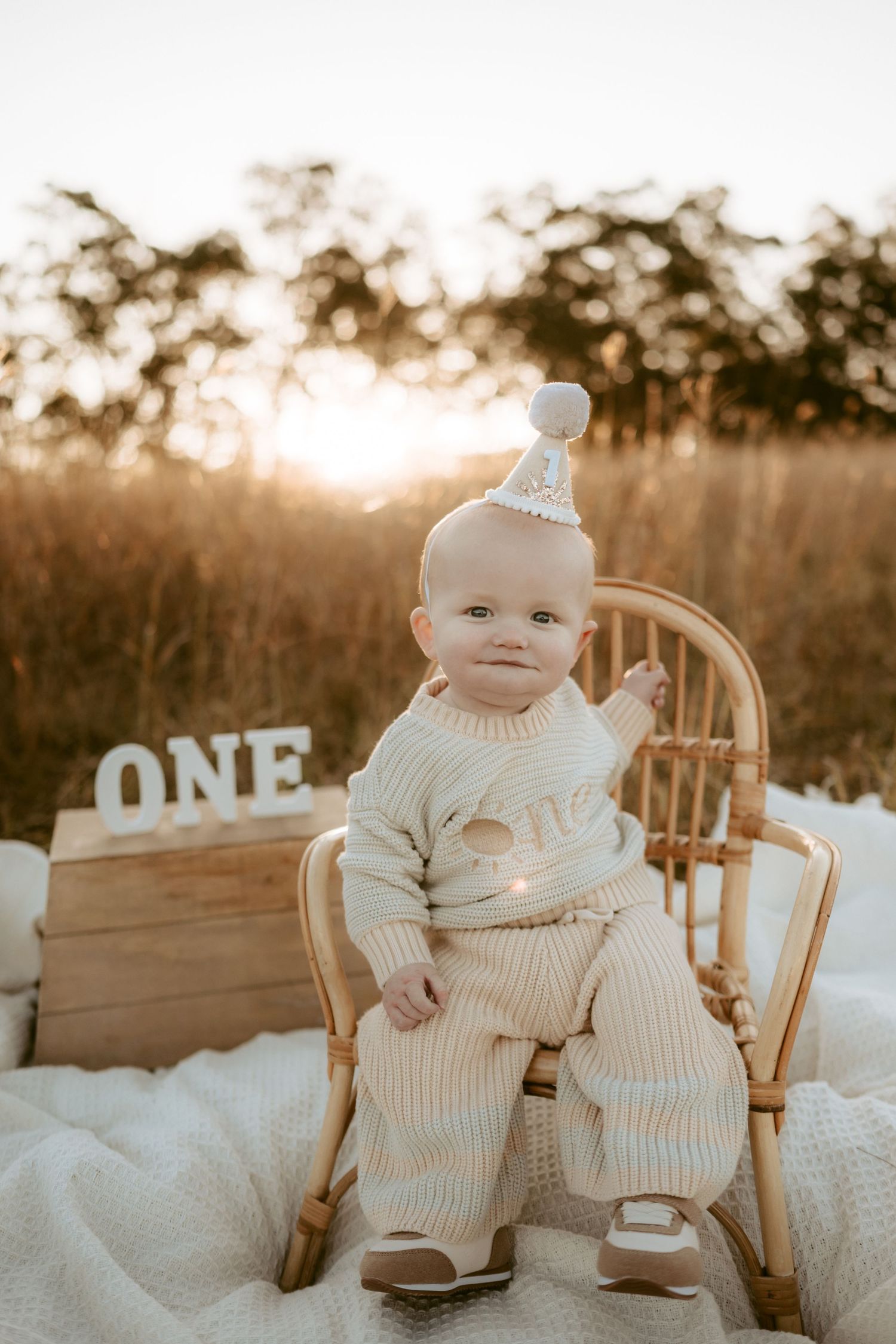 Baby wearing white knit outfit sits in wooden chair during golden hour photoshoot.
