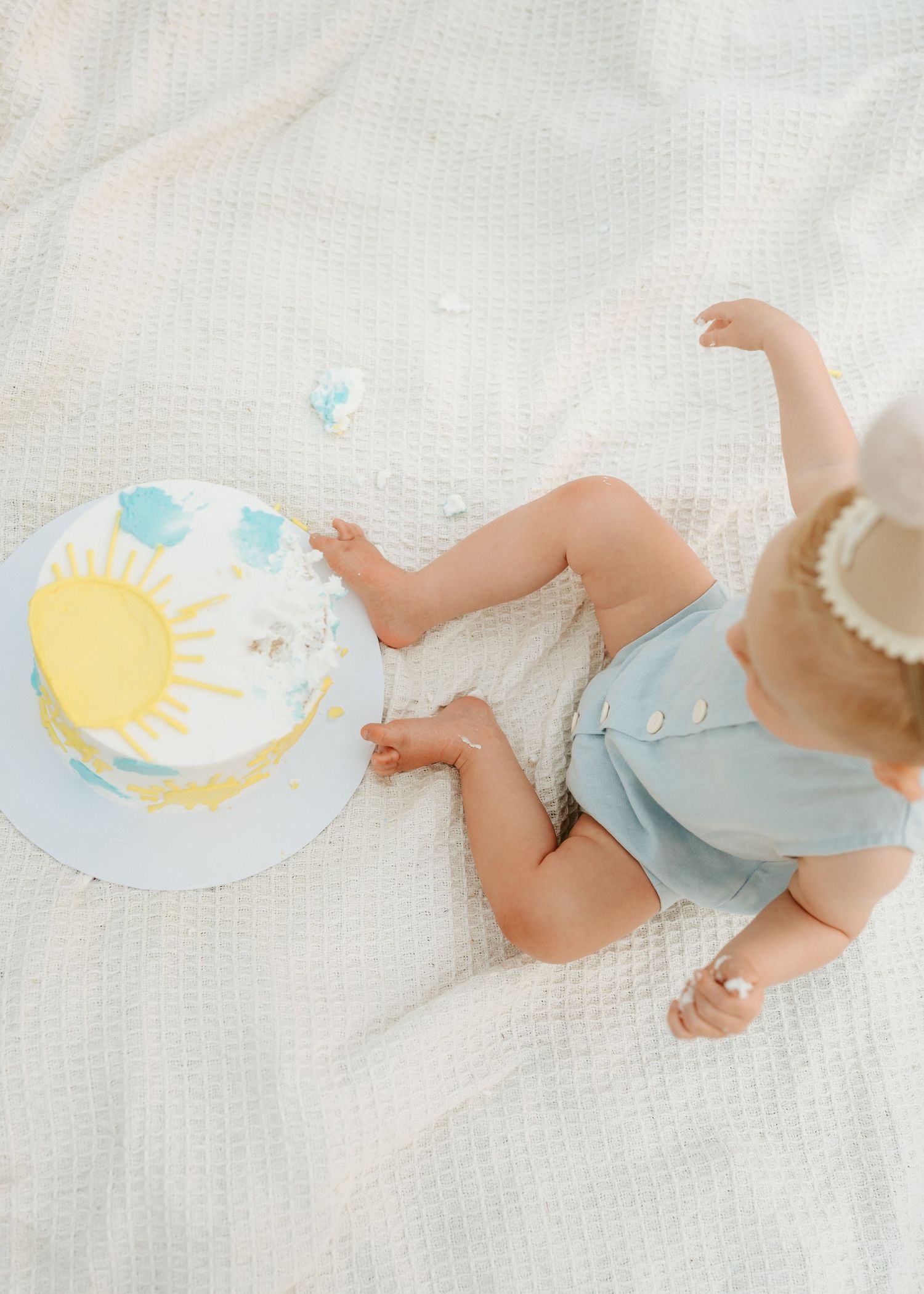 Baby in light blue outfit sitting on white textured blanket with sun cake.