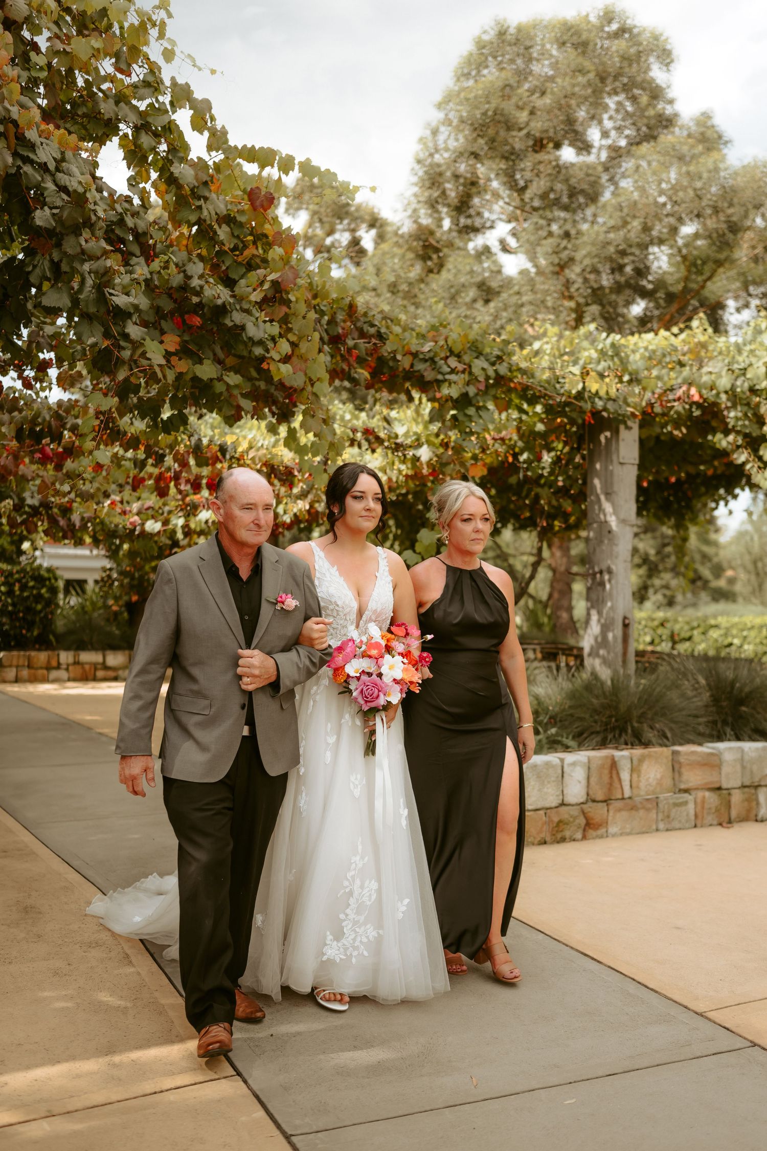 Wedding party walking together on a stone path under vine-covered pergola.
