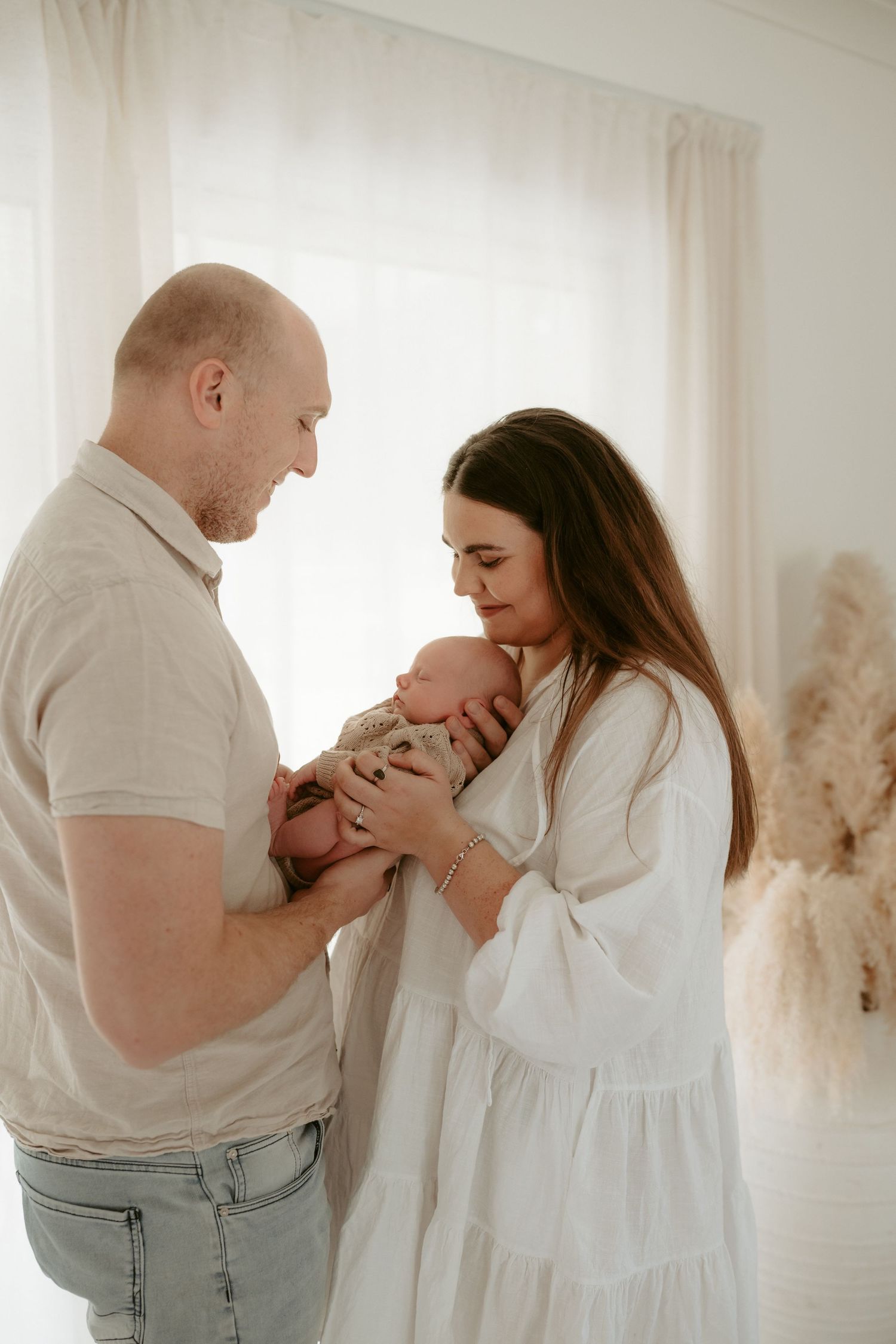 Parents in white and beige clothing tenderly holding their newborn in a bright minimalist room.