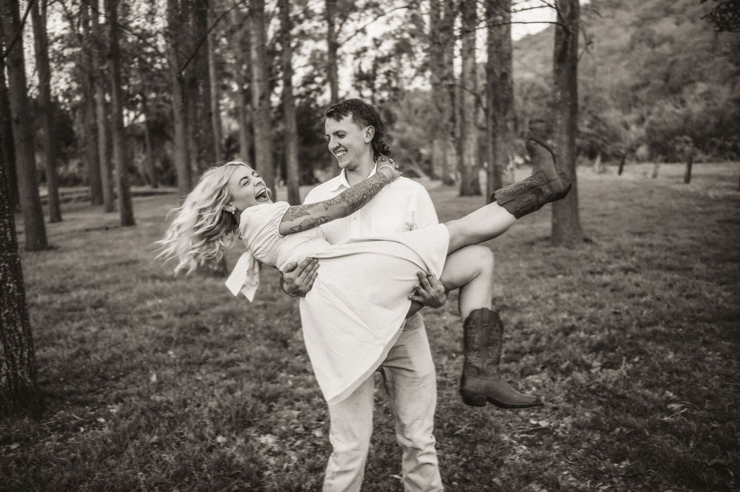 A romantic couple embraces playfully in a wooded park setting.