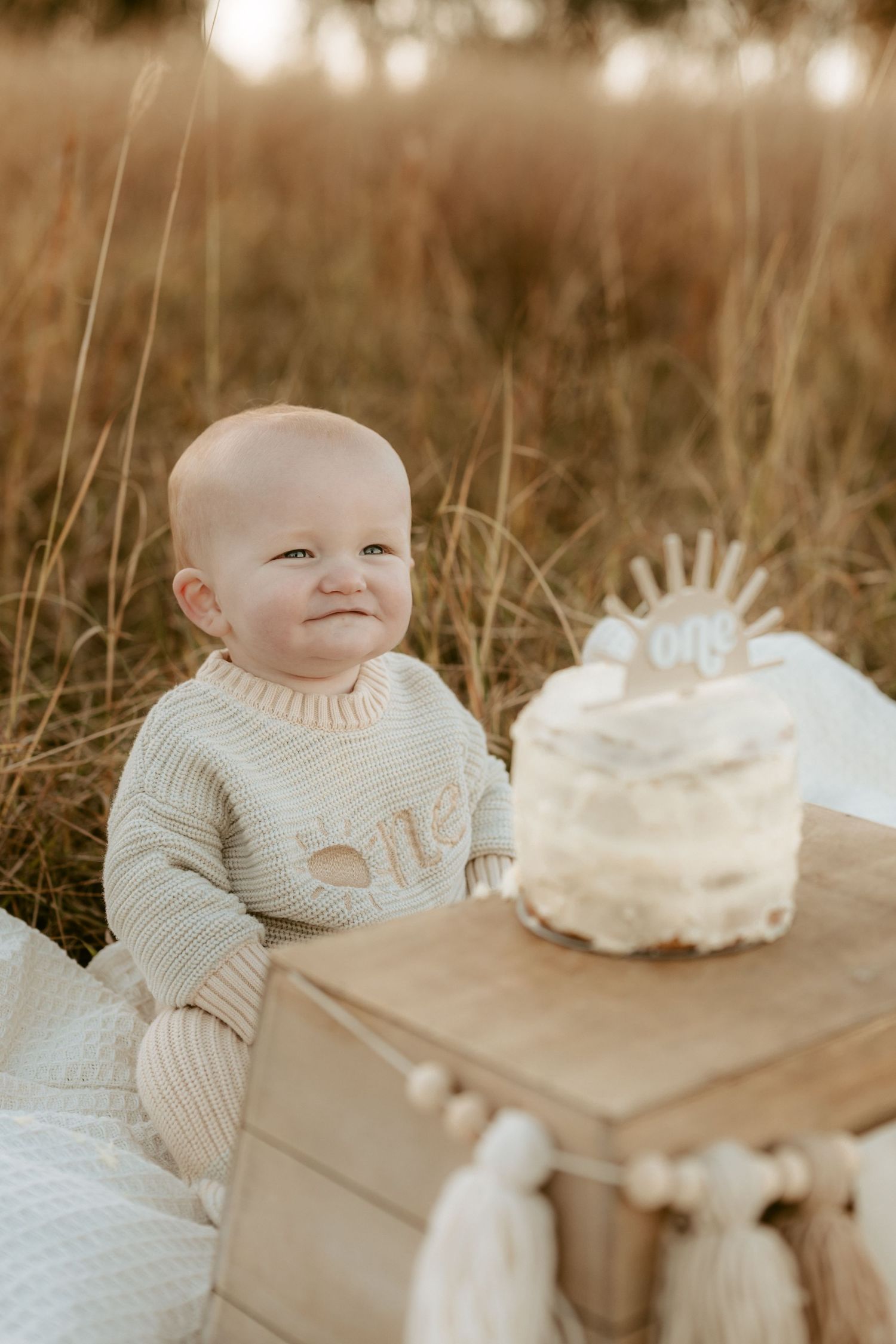 Baby in cream sweater smiles near rustic cake setup during outdoor photoshoot.