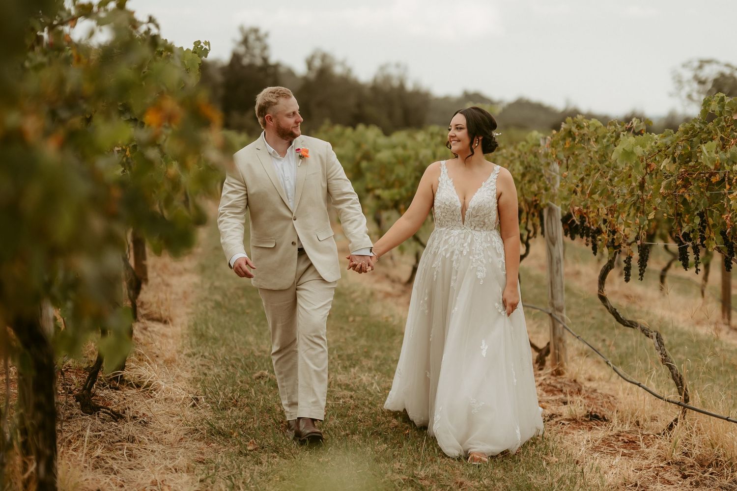 A couple in light colored wedding attire walks hand in hand through a scenic vineyard on a sunny afternoon.