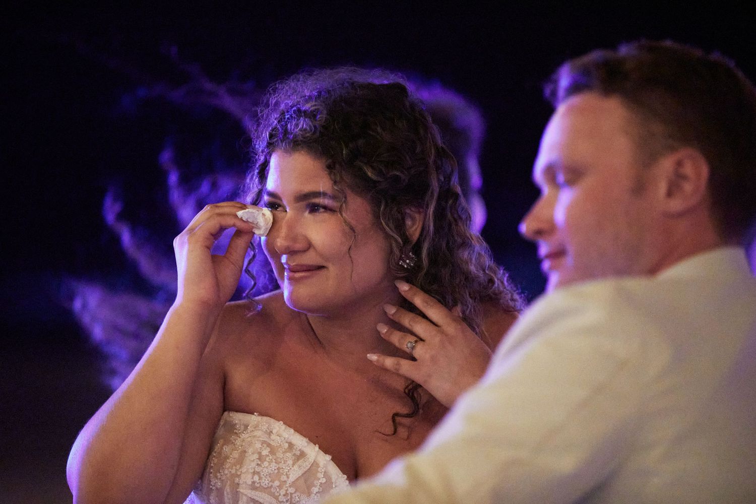 Bride wiping tears away listening to heartfelt speeches as husband looks on.