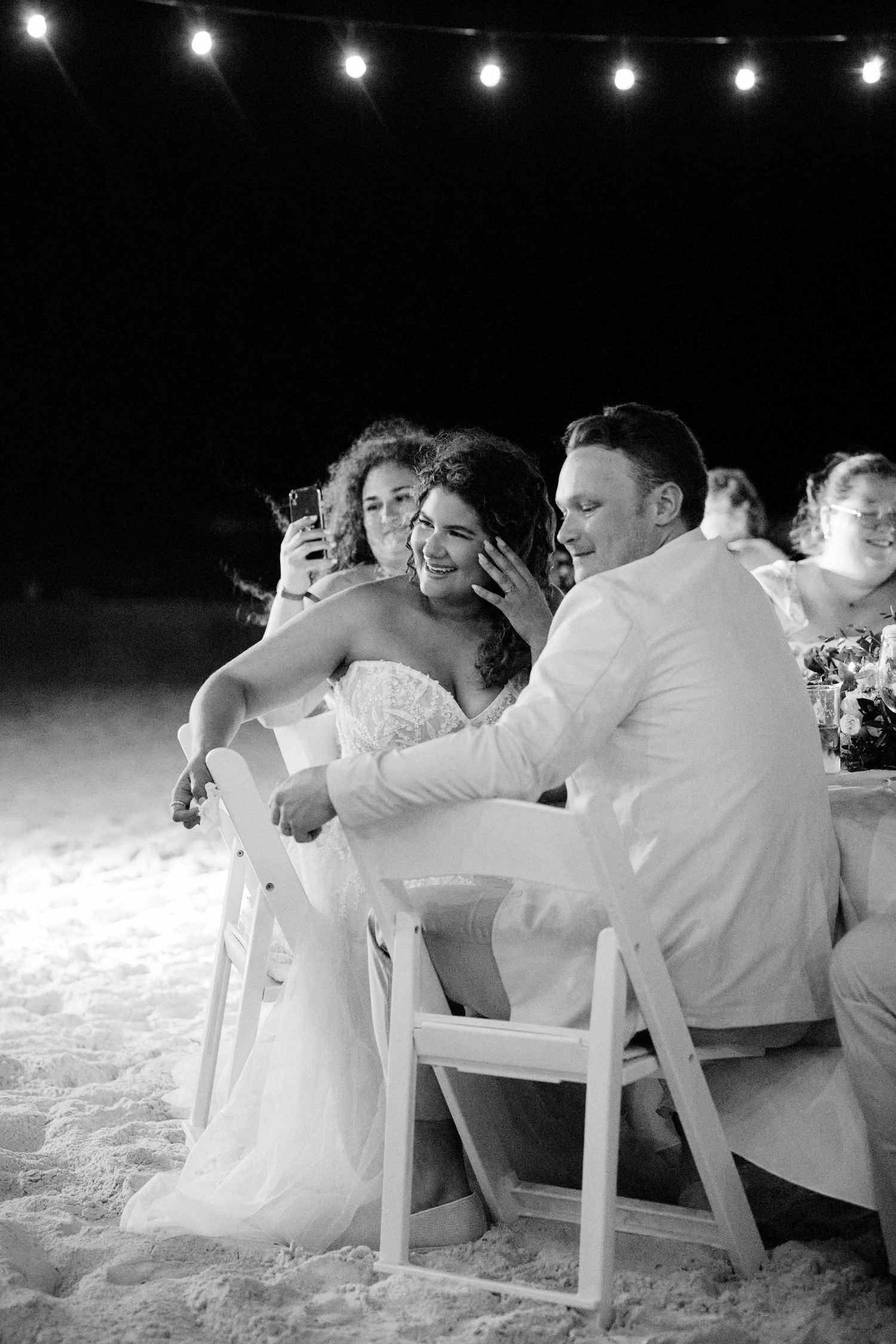 Bride and groom laughing and listening to heartfelt speeches as husband looks on.