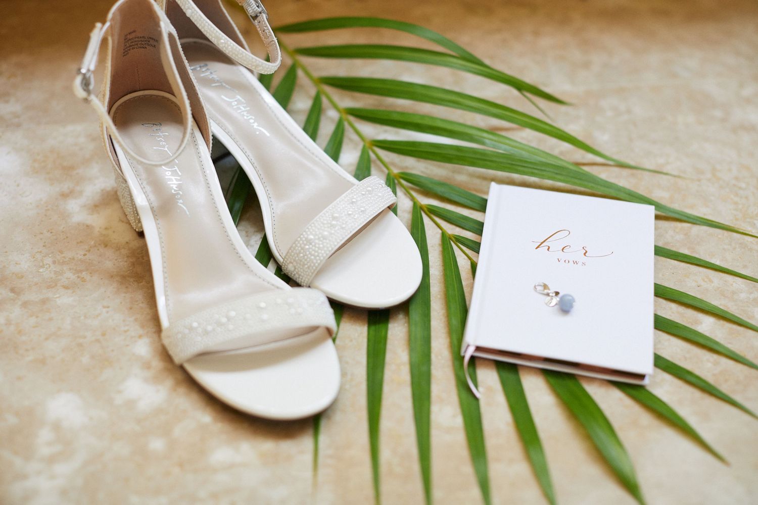 Brides wedding shoes, charm and vow book sitting on a tropical leaf.