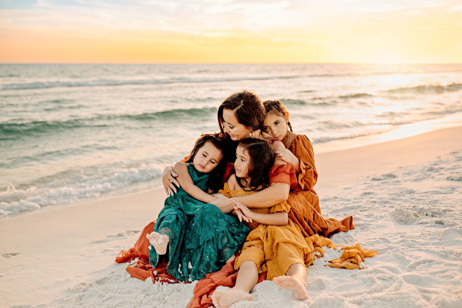 A mother and her 3 daughters sitting on the sand at sunset, embracing each other in a warm, loving hug, with the soft glow of the sunset.