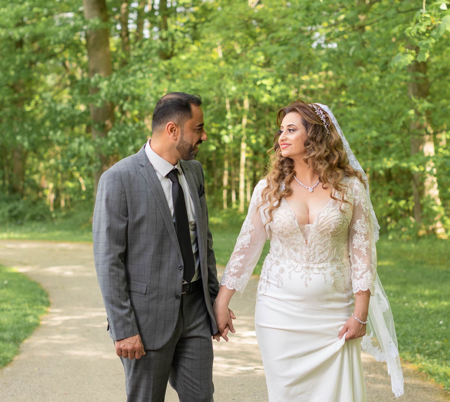 A couple in wedding attire walks together on a tree-lined path during a romantic outdoor photo session.