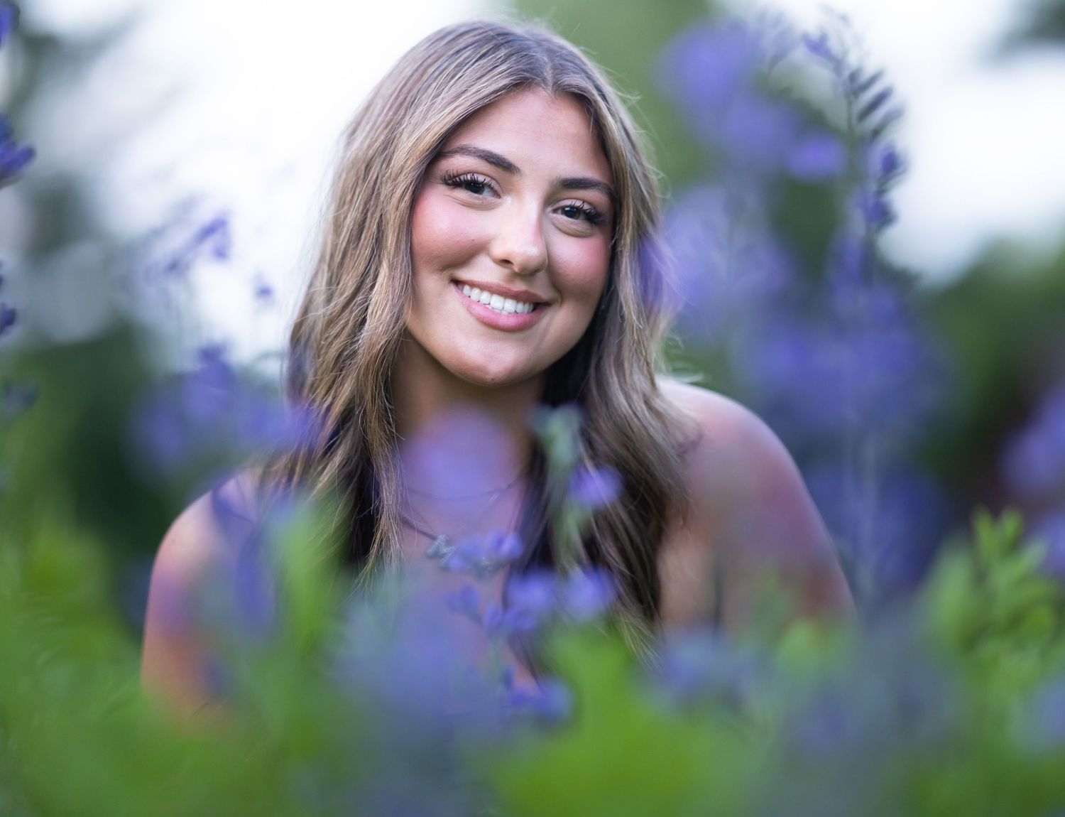 Young woman with long hair smiling among purple wildflowers in a soft-focus outdoor portrait.
