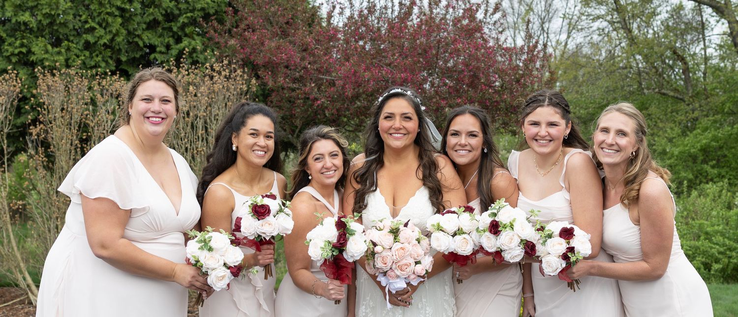 Wedding party in white dresses holding bouquets poses together outdoors in a garden setting.