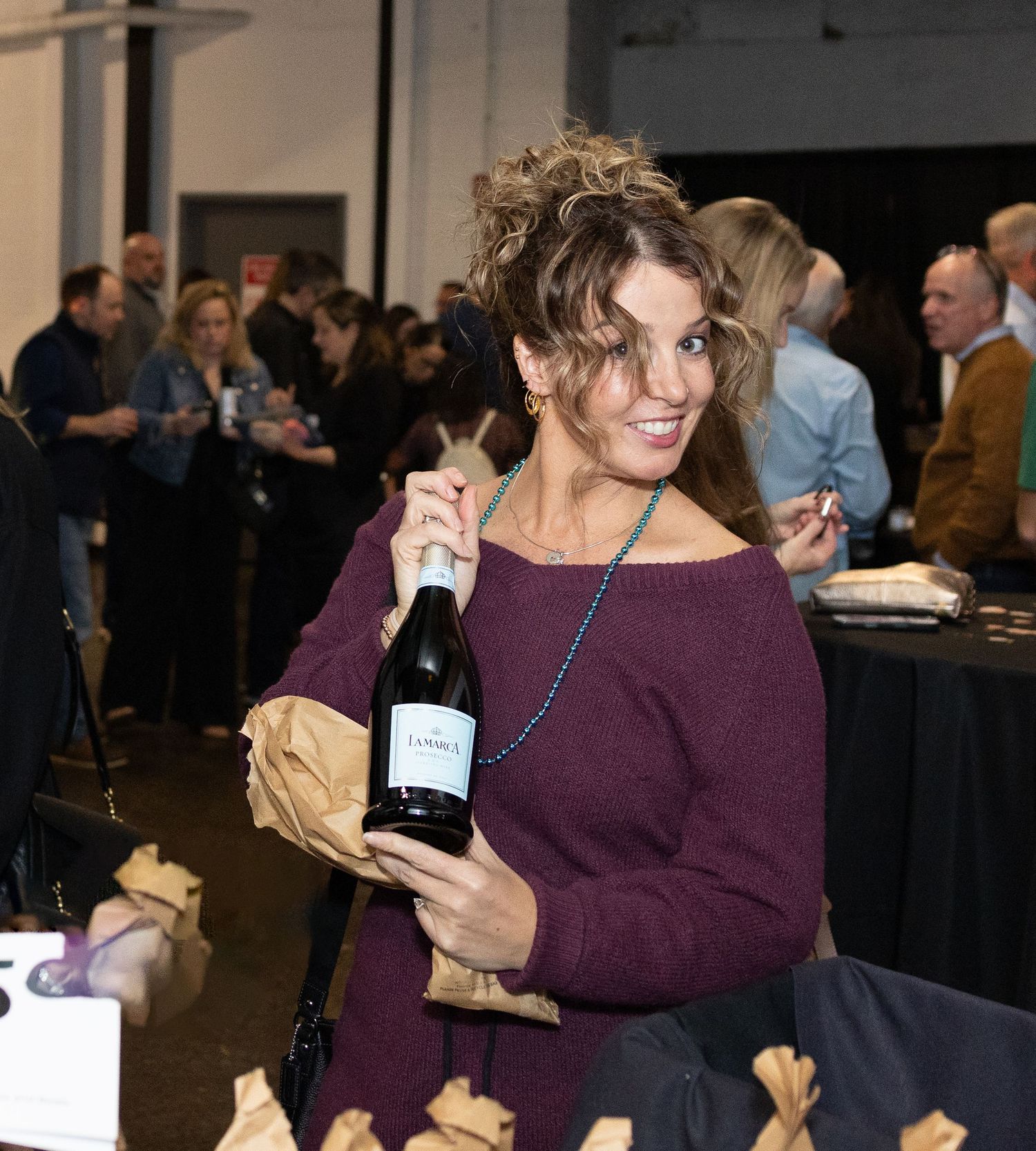 Someone in a purple sweater holds a wine bottle and brown paper bag at a social gathering.