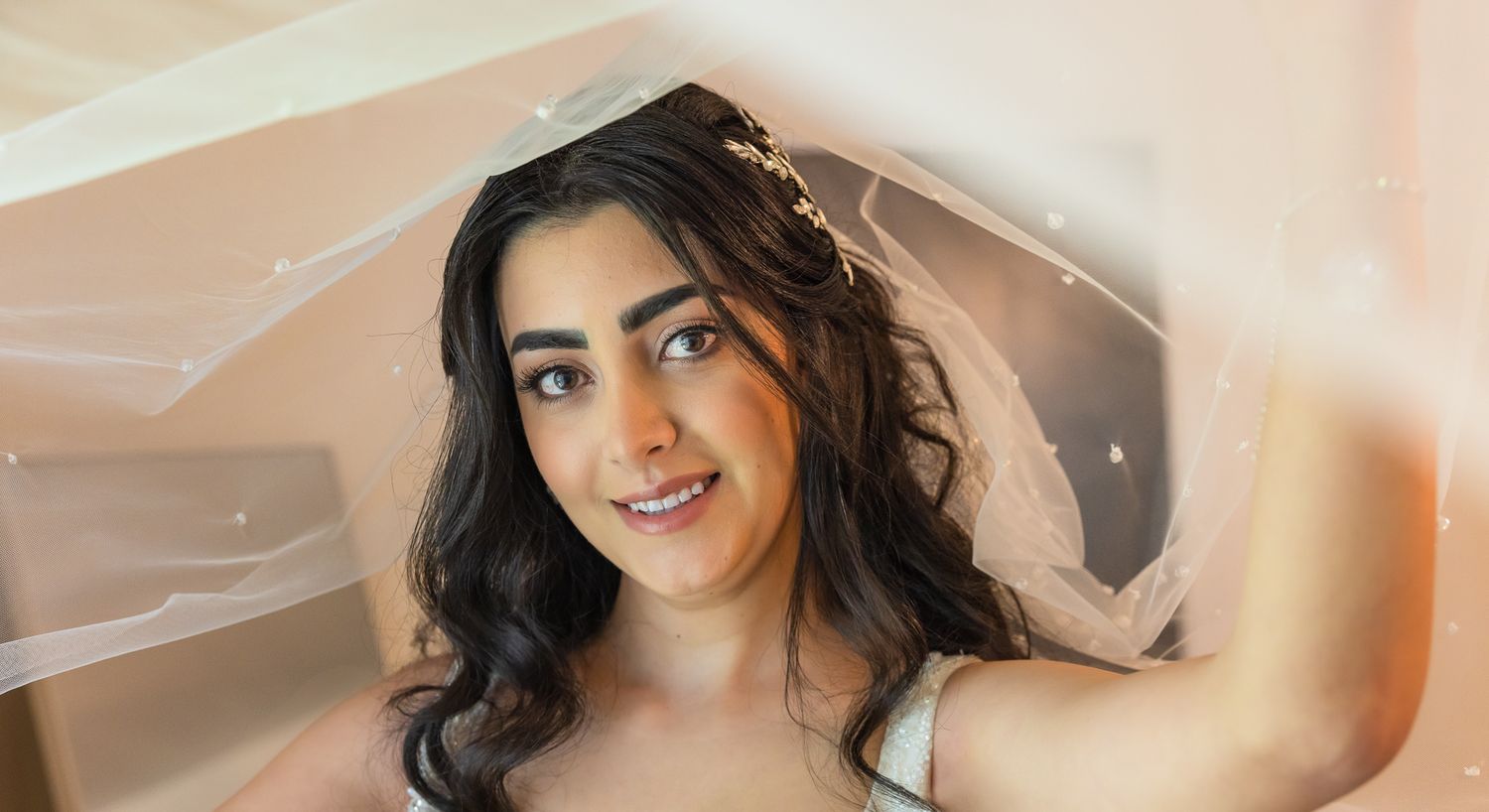 A smiling person wearing a veil poses in soft peachy lighting for a bridal portrait.