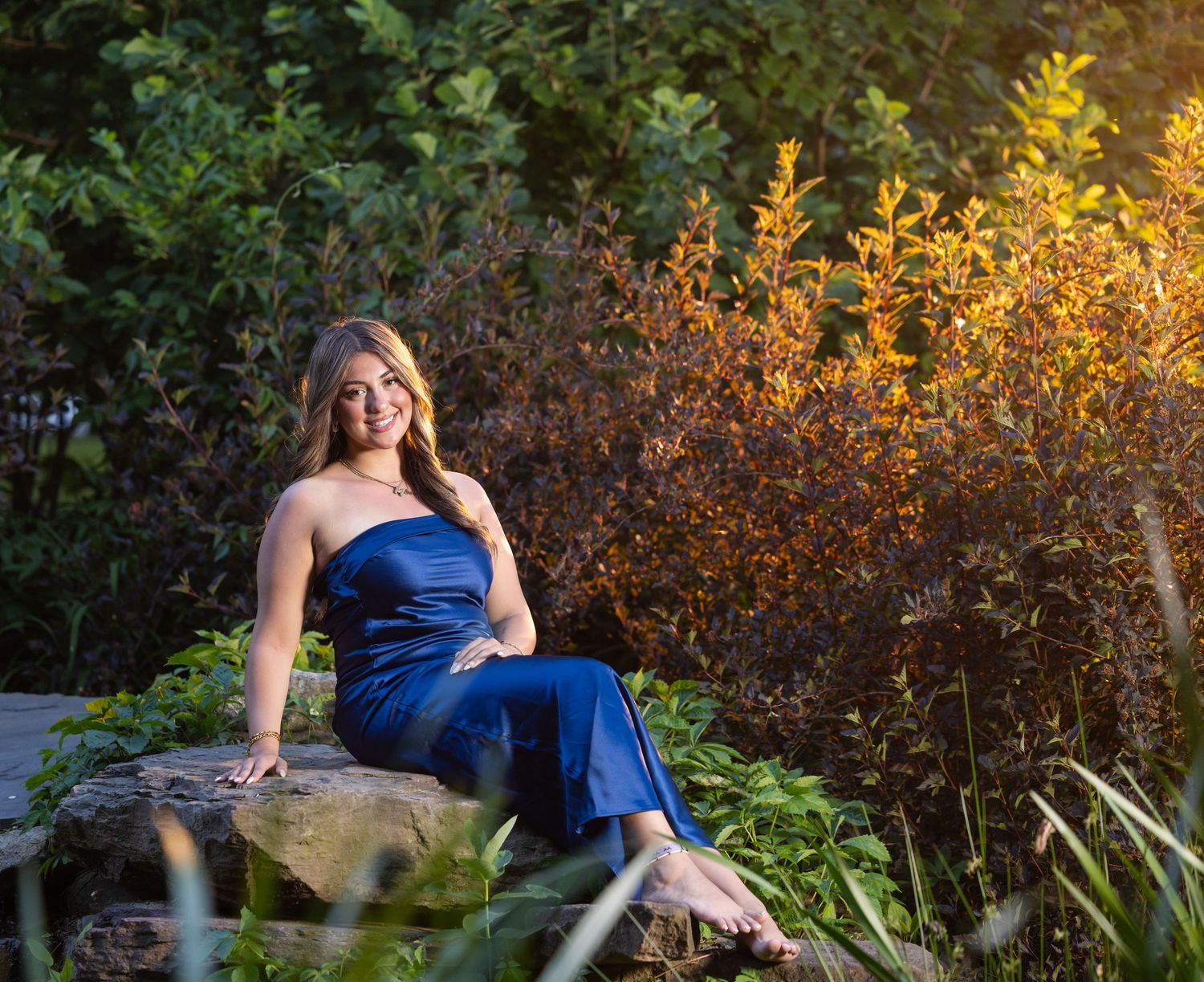 Young woman in blue jumpsuit sitting on rocks surrounded by lush green and autumn foliage in golden hour light.