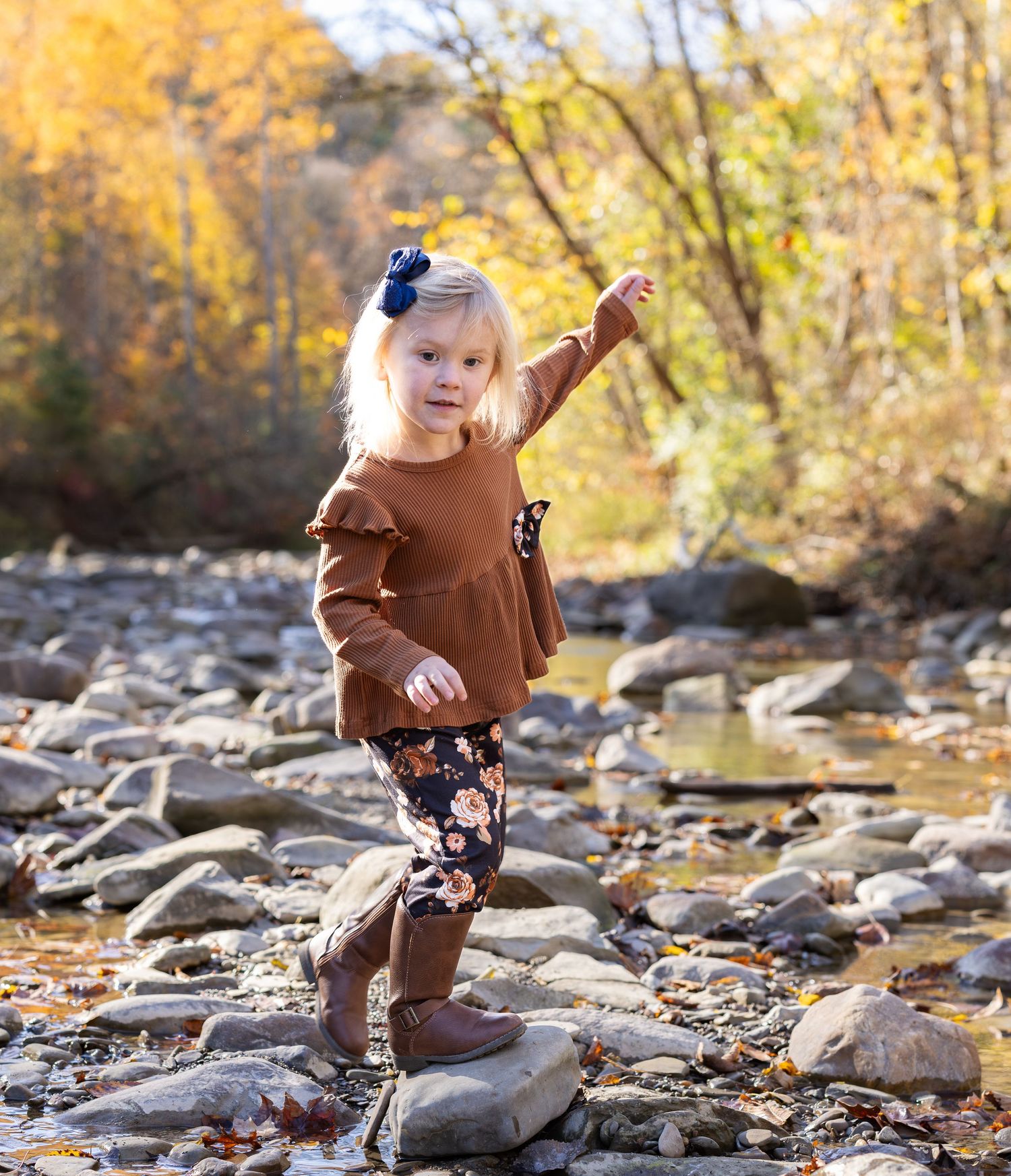 Child in brown sweater plays on rocky riverbank surrounded by golden autumn foliage.