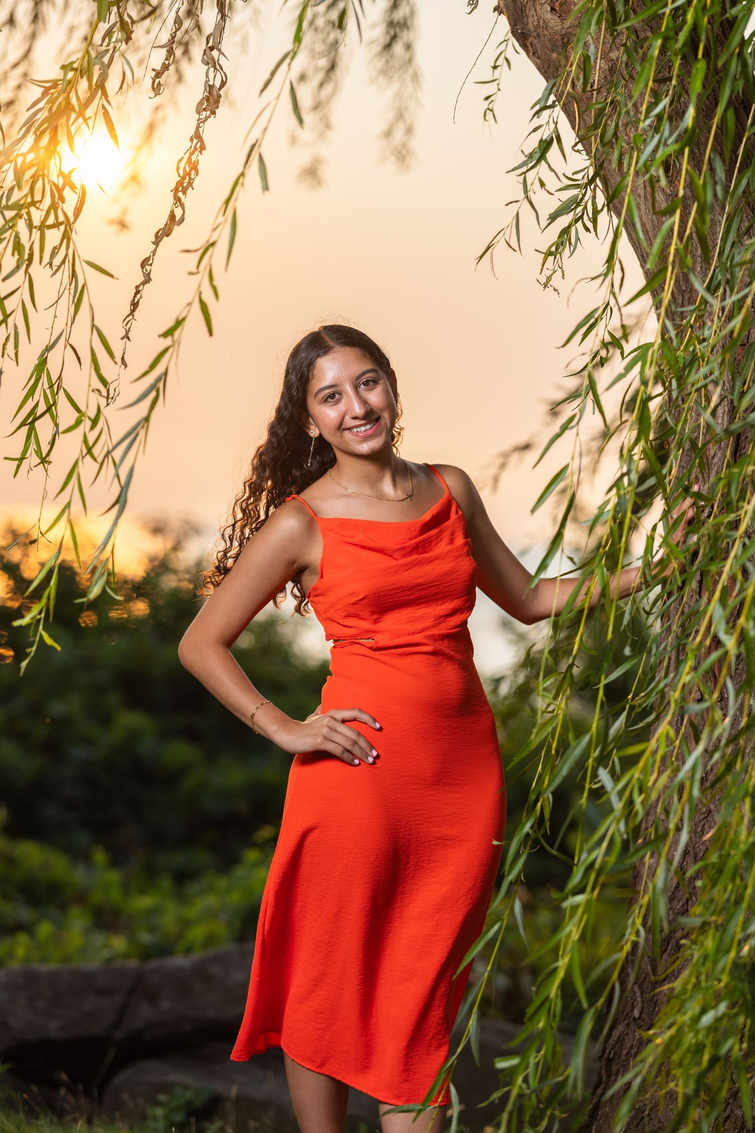 A person in a bright red dress poses under a willow tree at sunset creating a warm summer portrait.