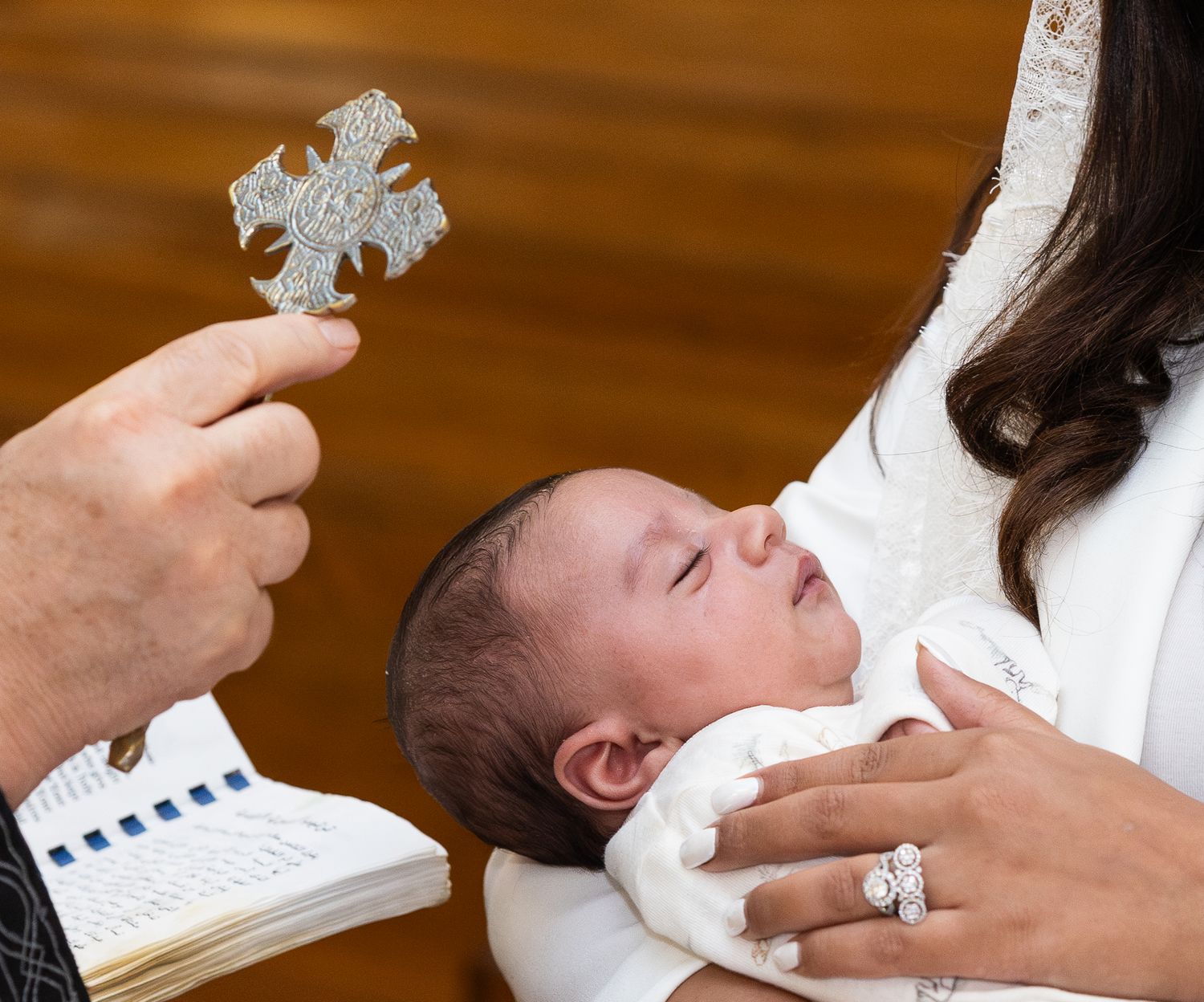 A small infant in white clothing is held during a baptism ceremony with a cross visible in the foreground.