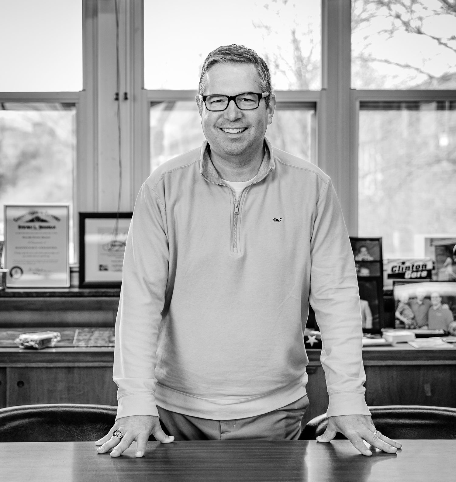 Person in glasses and white pullover smiling while leaning on desk in bright office space.