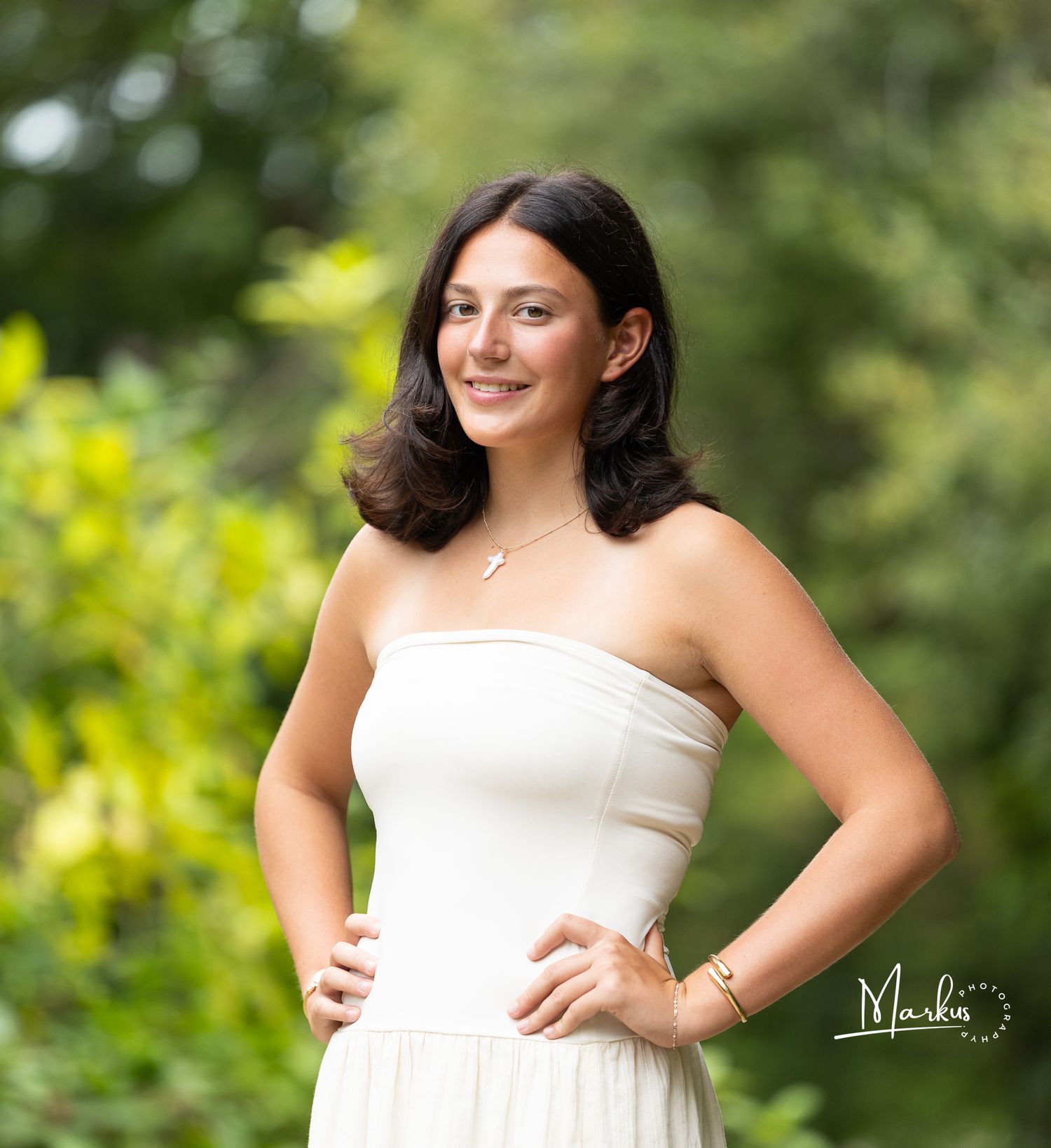 Young woman in white strapless dress smiling outdoors with lush green foliage background, professional portrait photo.