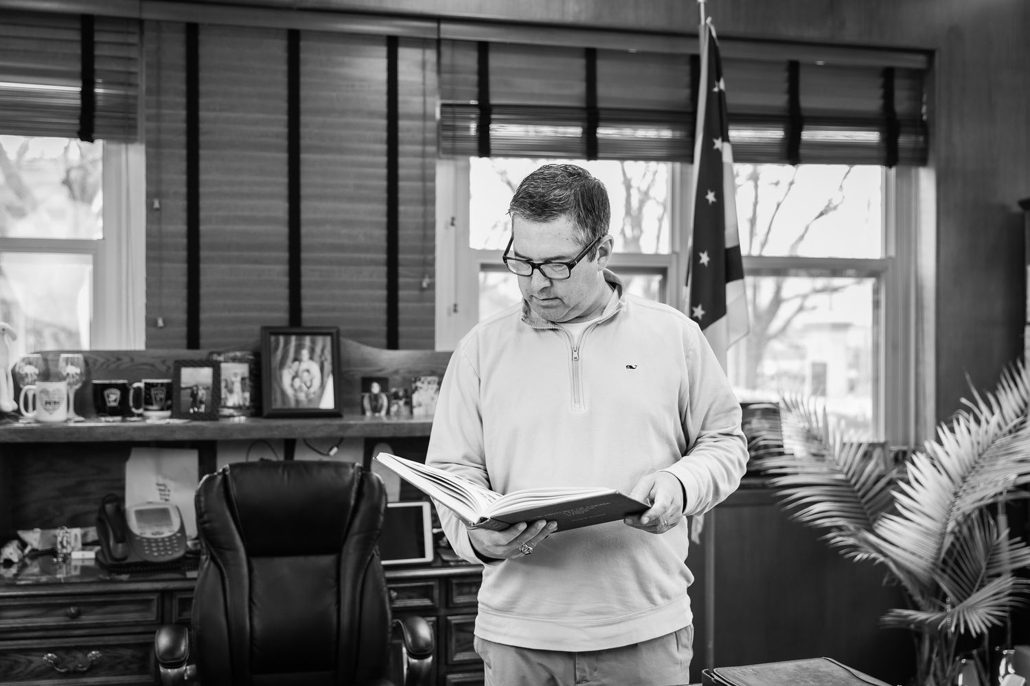 A black and white photograph shows a person reading a book while seated in a cozy interior setting.