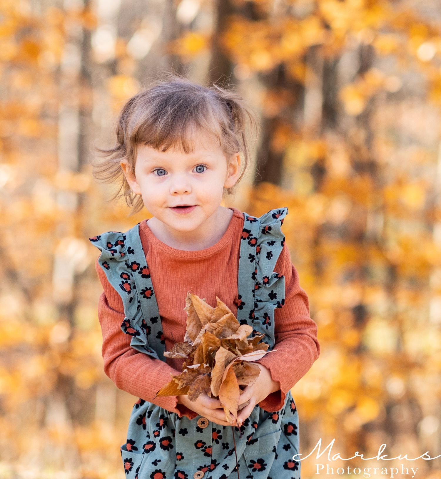 A small child wearing a floral dress and orange shirt plays with autumn leaves in a golden outdoor setting.