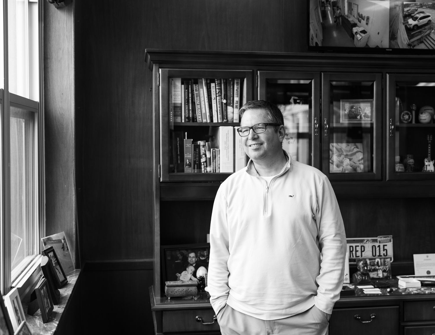 Black and white photo of person in glasses standing in home office with bookshelf and window.