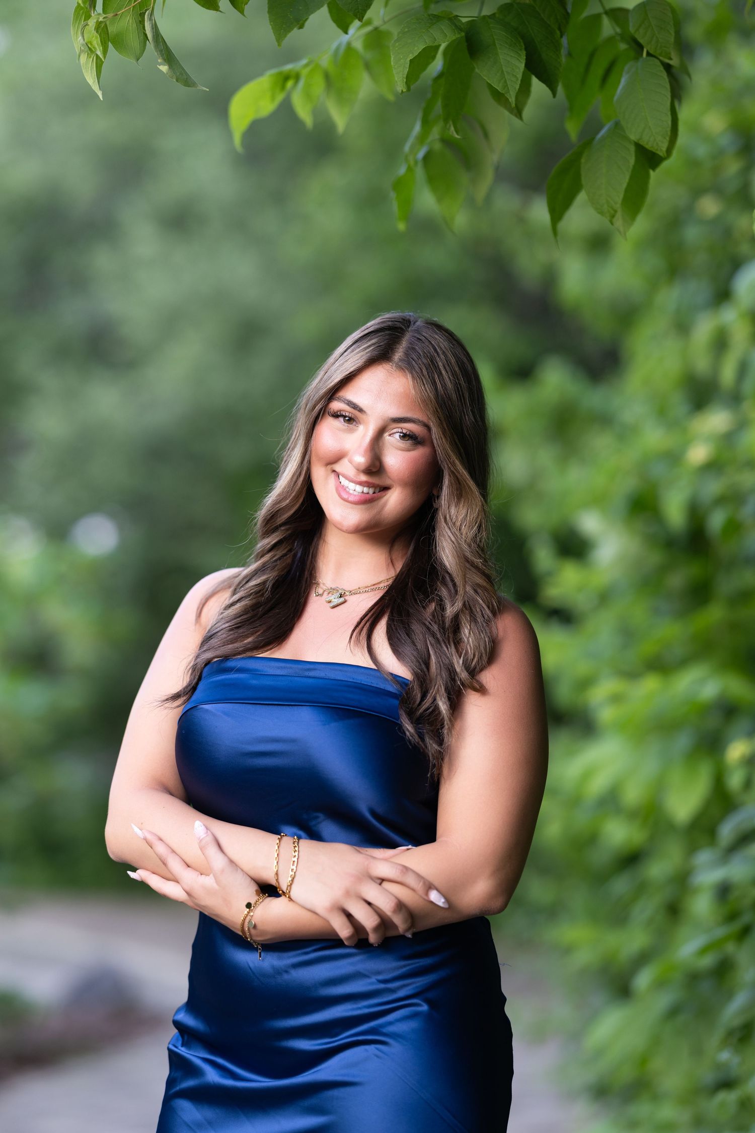 Young woman in blue satin strapless dress smiling outdoors, wearing gold jewelry, with lush green foliage background.