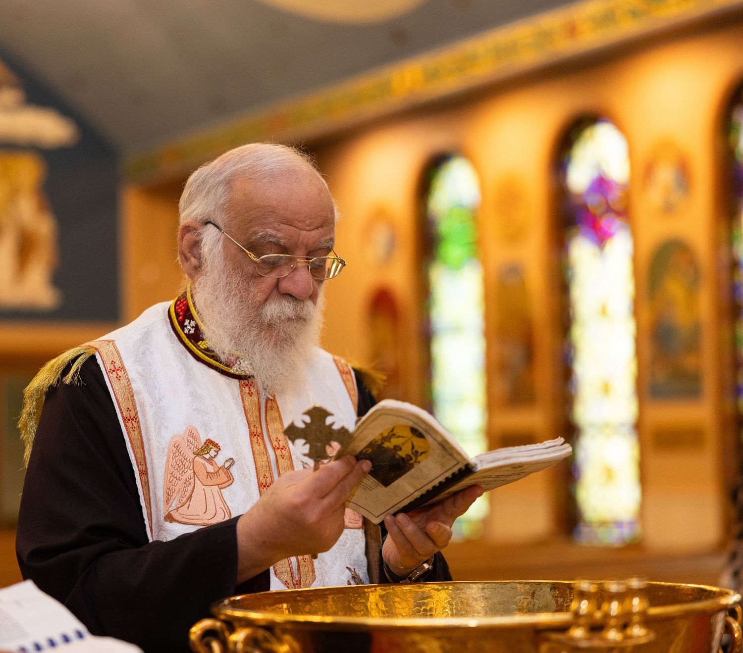 An elderly man with a white beard lights a candle in an ornate church with stained glass windows in the background.