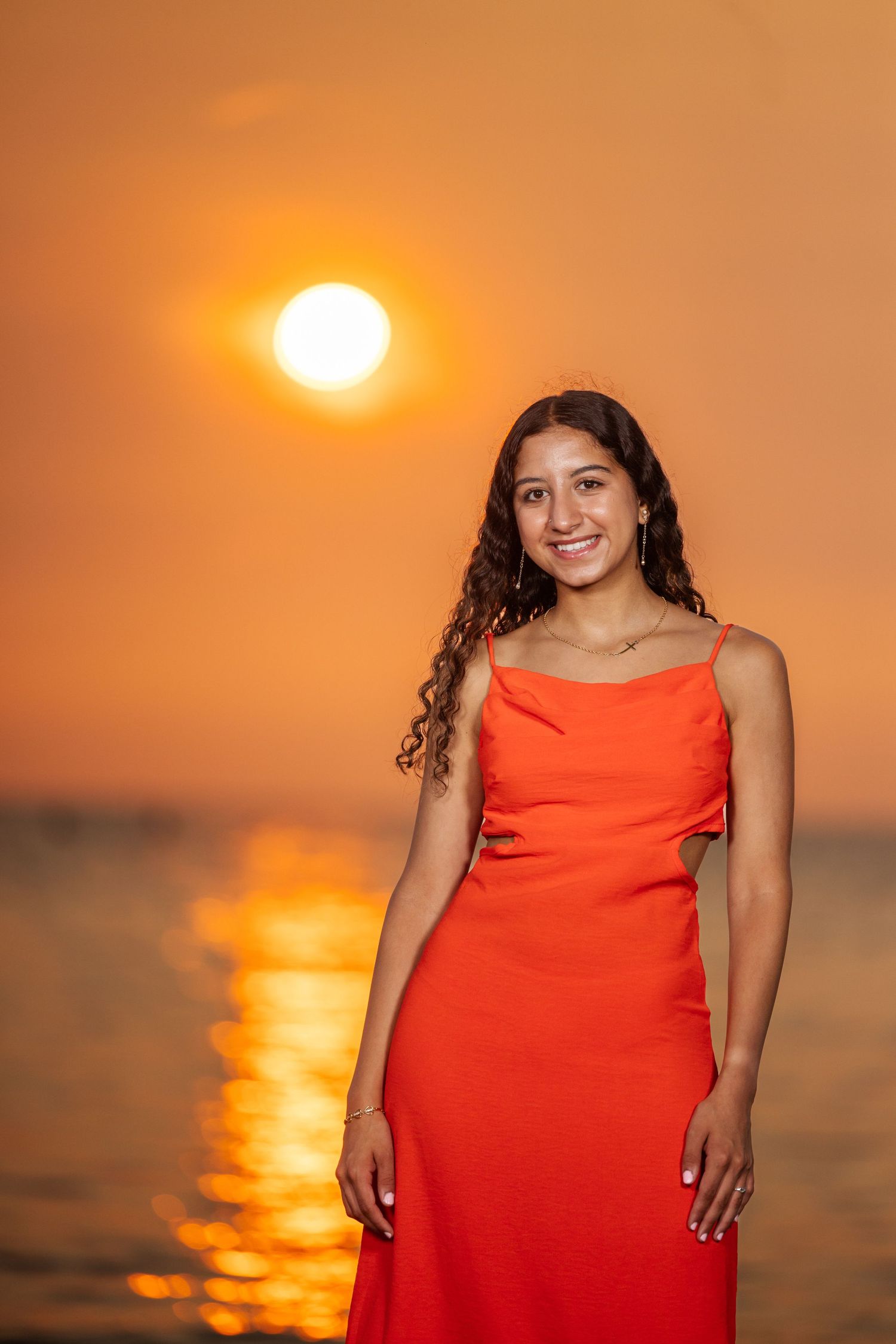 A person in a vibrant red dress poses against a stunning orange sunset over calm ocean waters.
