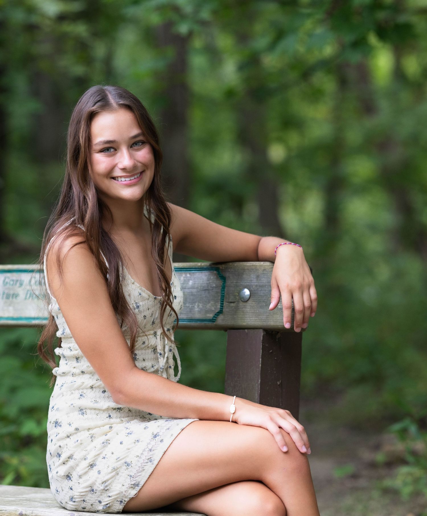 A person in a floral dress sits on a wooden bench outdoors with a green forest backdrop on a sunny day.