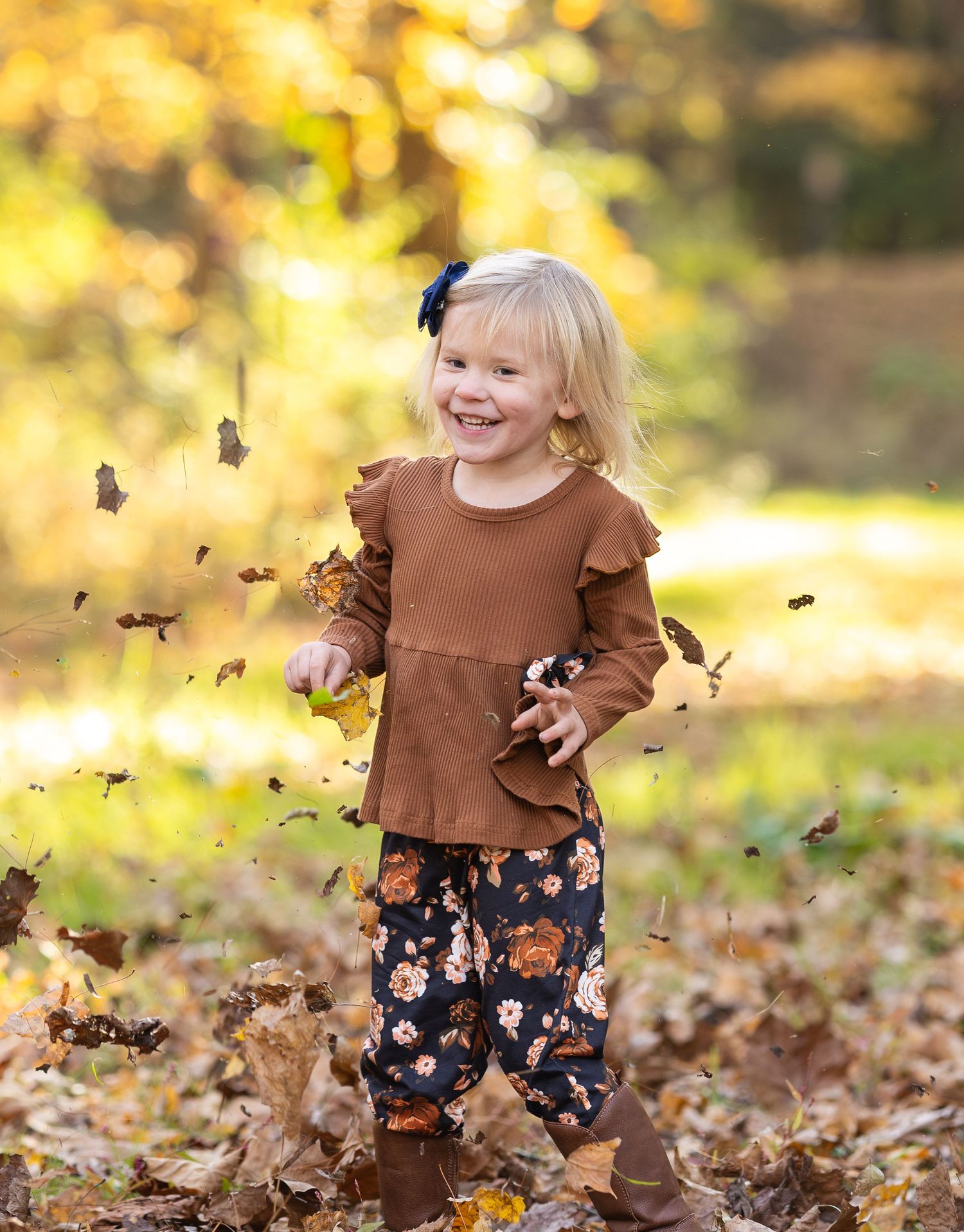 A child in a brown sweater and floral dress plays joyfully in a pile of autumn leaves on a sunny fall day.