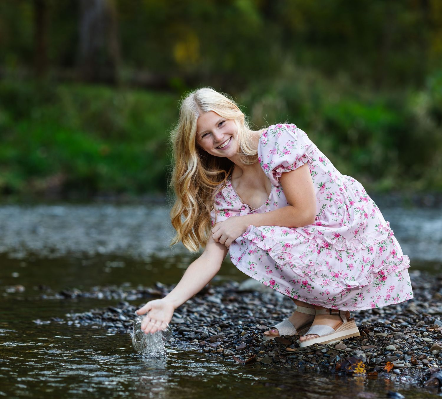 Woman in floral dress plays and splashes in shallow water along rocky riverbank on summer day.