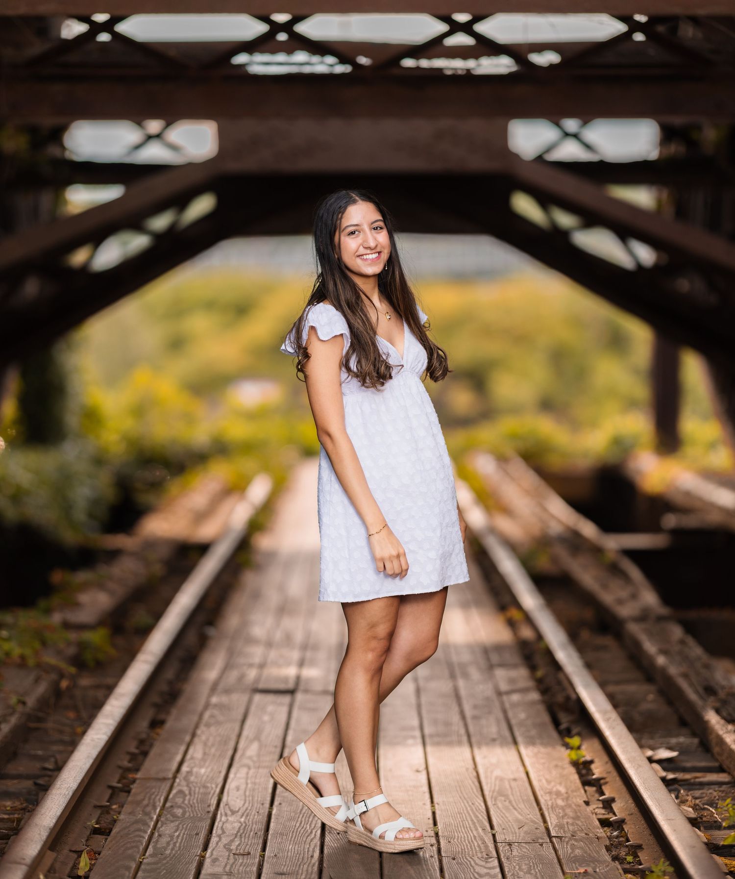 A person in a white dress poses on wooden railroad tracks surrounded by autumn foliage at sunset.